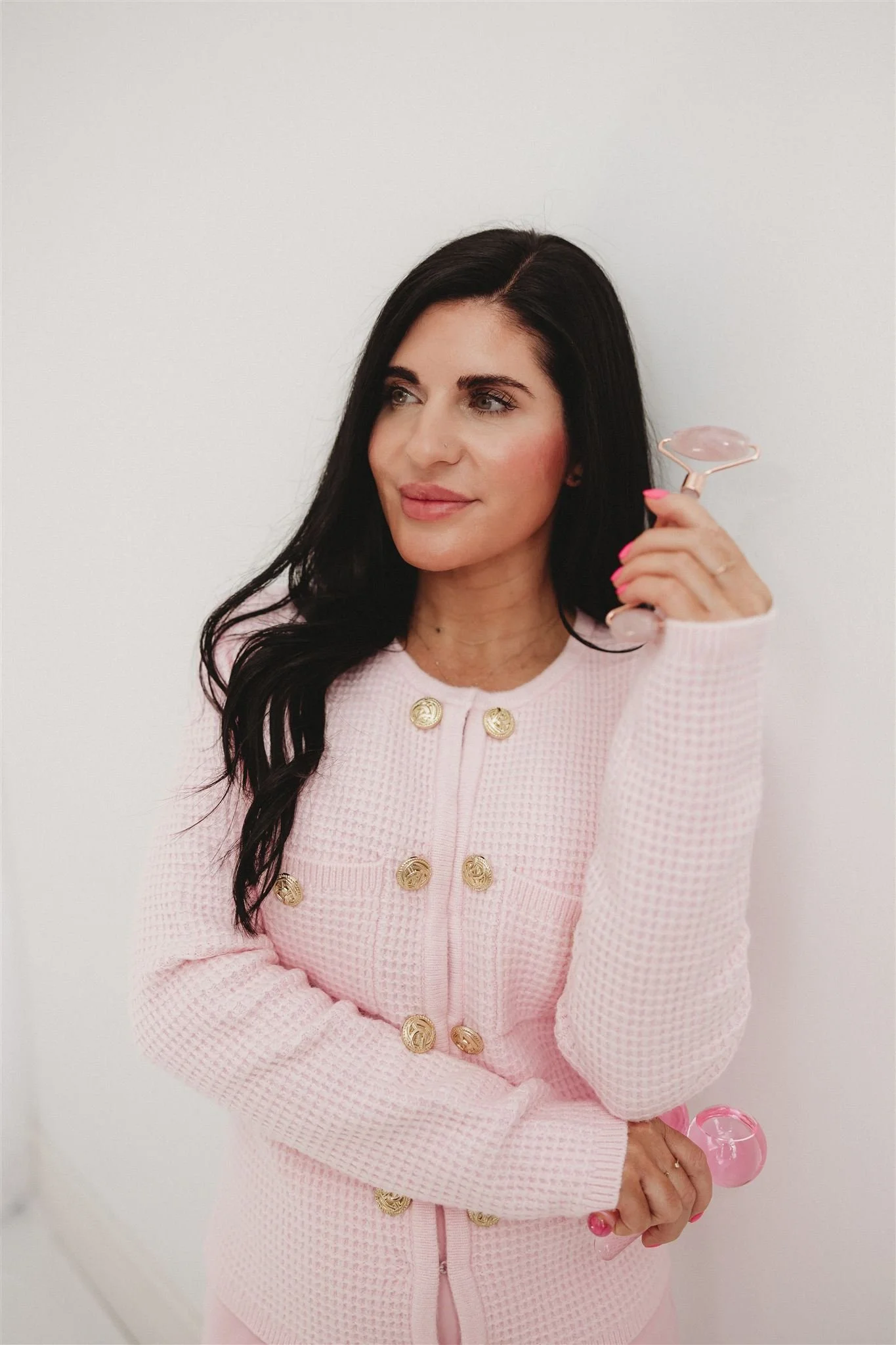 Woman with long dark hair holding a pink facial roller, wearing a pink textured jacket with gold buttons, standing against a plain white wall.