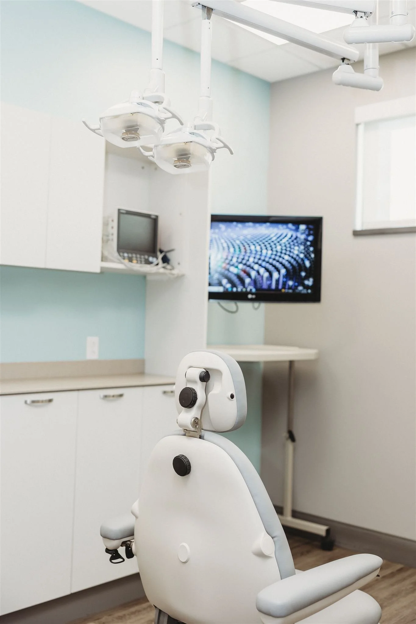 Dental examination room with dental chair, overhead dental lights, medical monitor, and a wall-mounted TV.