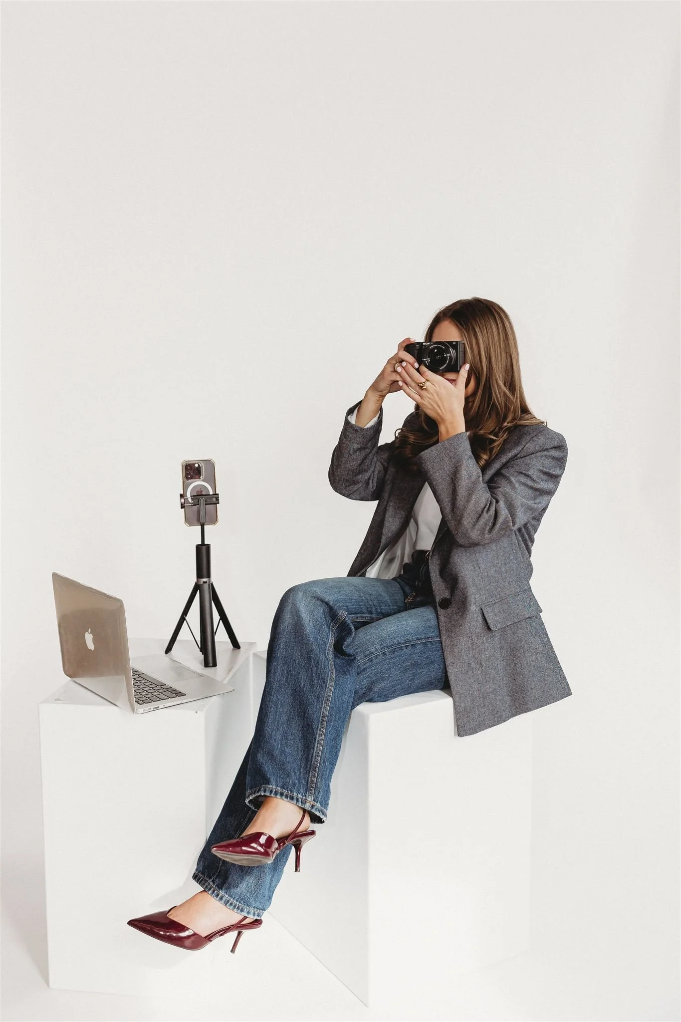A woman sitting on a white block, taking a photograph with a black camera. She is wearing a gray blazer, blue jeans, and red high-heeled shoes. There is a laptop and a smartphone on a smaller white block beside her.