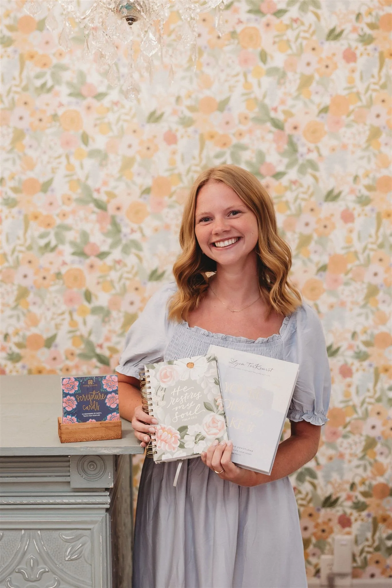 A woman smiling and holding a notebook and a book, standing in front of a floral wallpaper backdrop with a small table holding a box of gift cards.