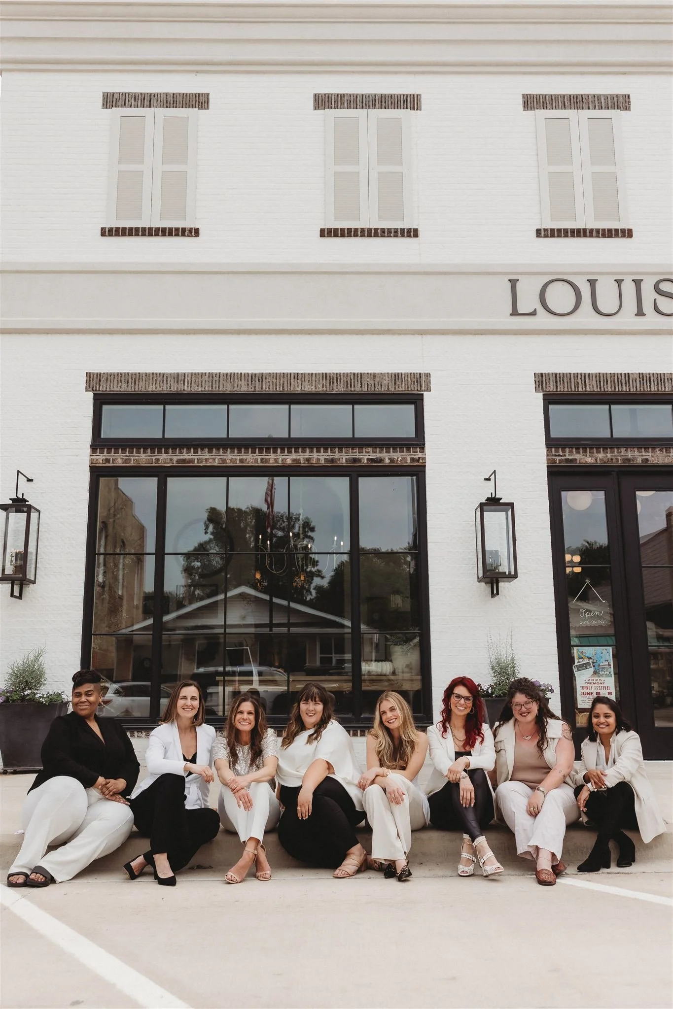 Group of eight women sitting on curb outside a building with large windows and a sign that reads 'LOUIS'