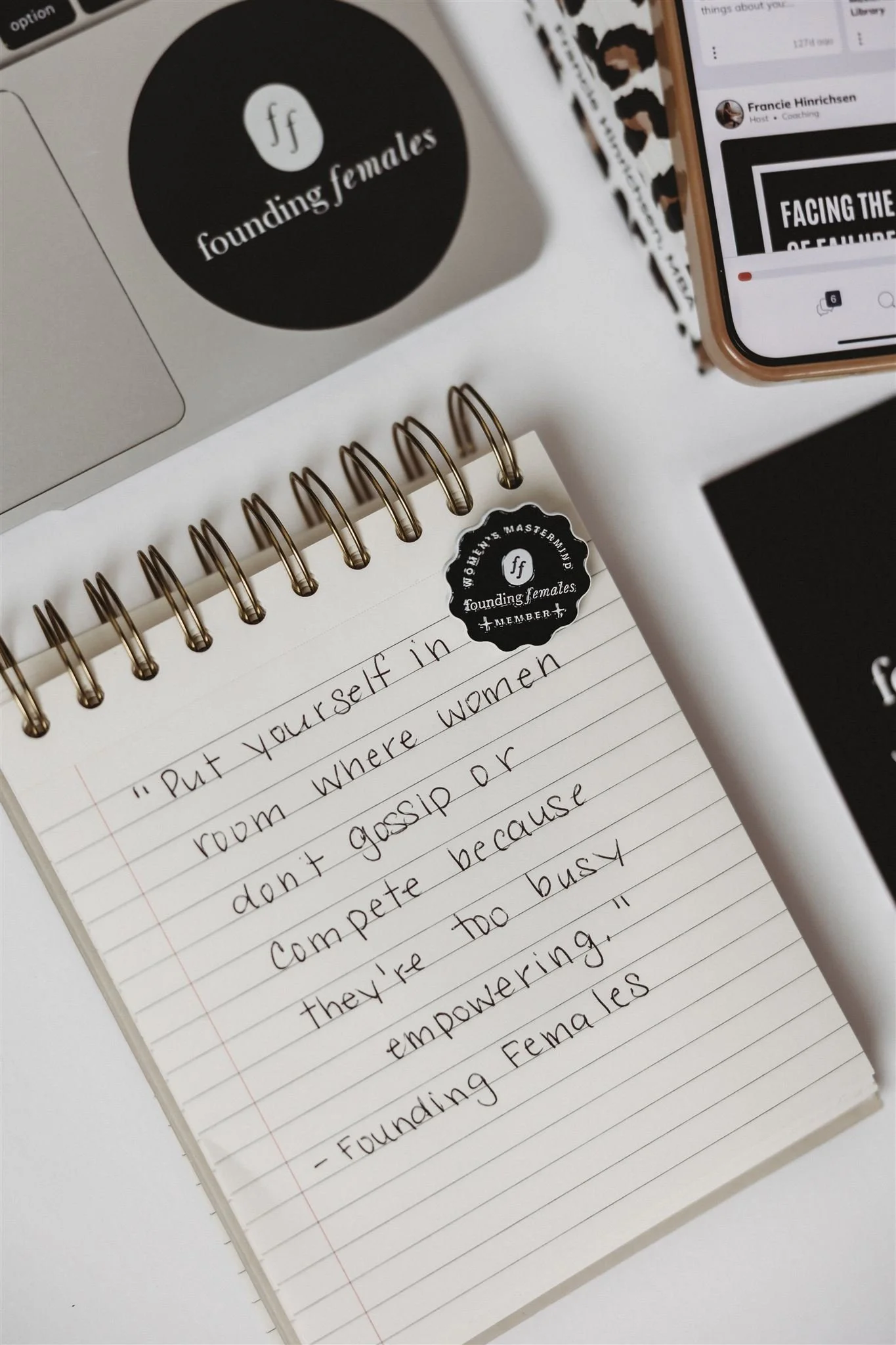 A spiral-bound notebook with a handwritten quote about women empowerment, a laptop with a founding females sticker, a phone with a social media page, and some other papers and stickers.