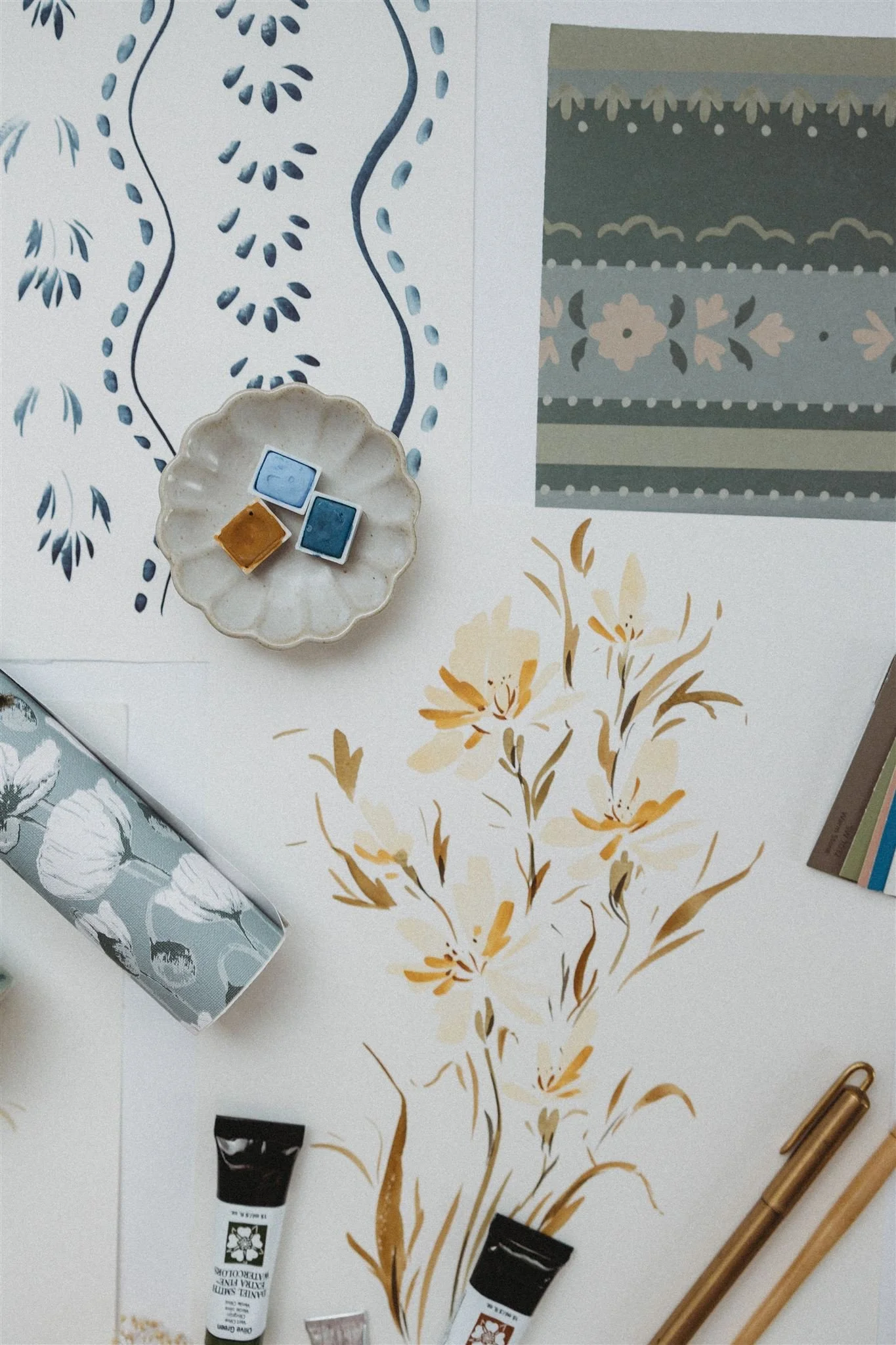 A flat lay of decorative paper, paint samples in a small dish, a patterned pencil case, and art supplies on a table.