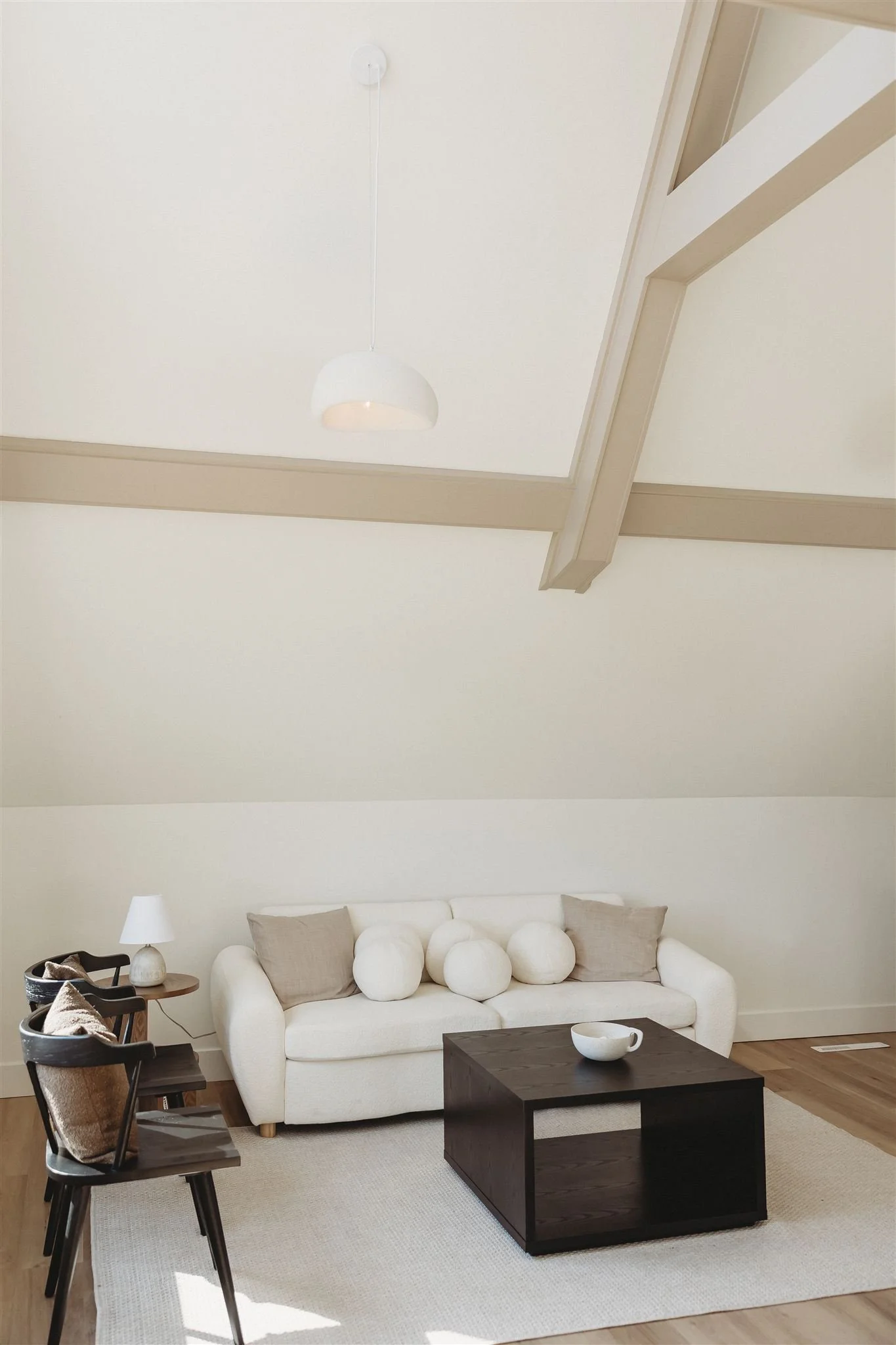 Living room with white sofa, beige cushions, dark wooden coffee table, side table with a lamp, two dark wooden chairs, white rug, and a ceiling light in a room with a sloped ceiling and light-colored walls.