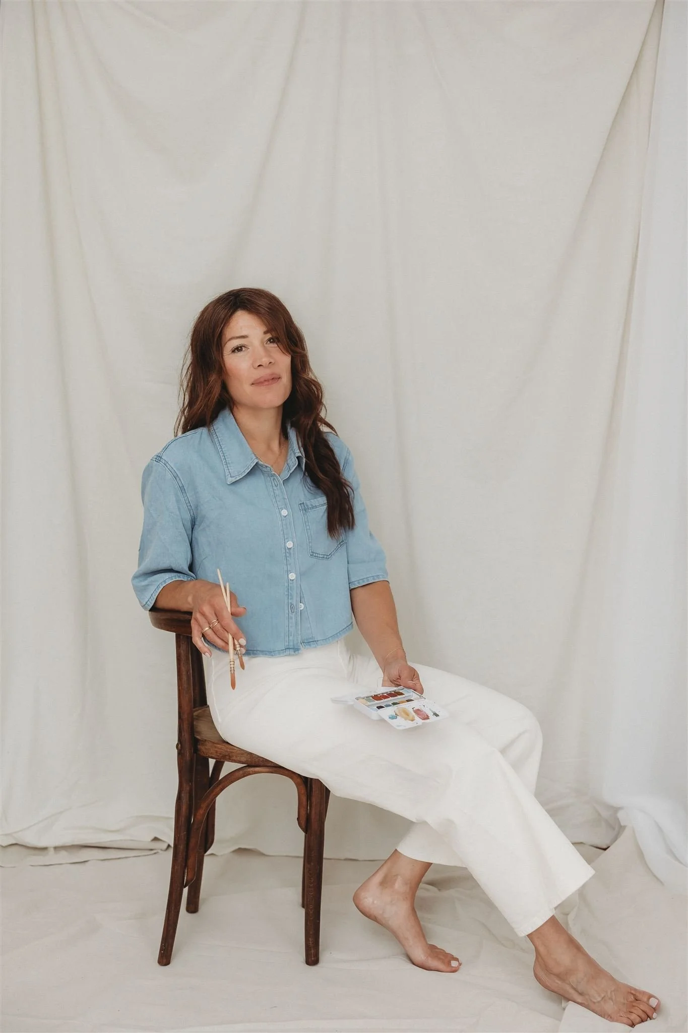 A woman with long wavy brown hair, wearing a light blue denim shirt and white pants, sitting on a wooden chair against a white backdrop, holding a paint palette and brushes.