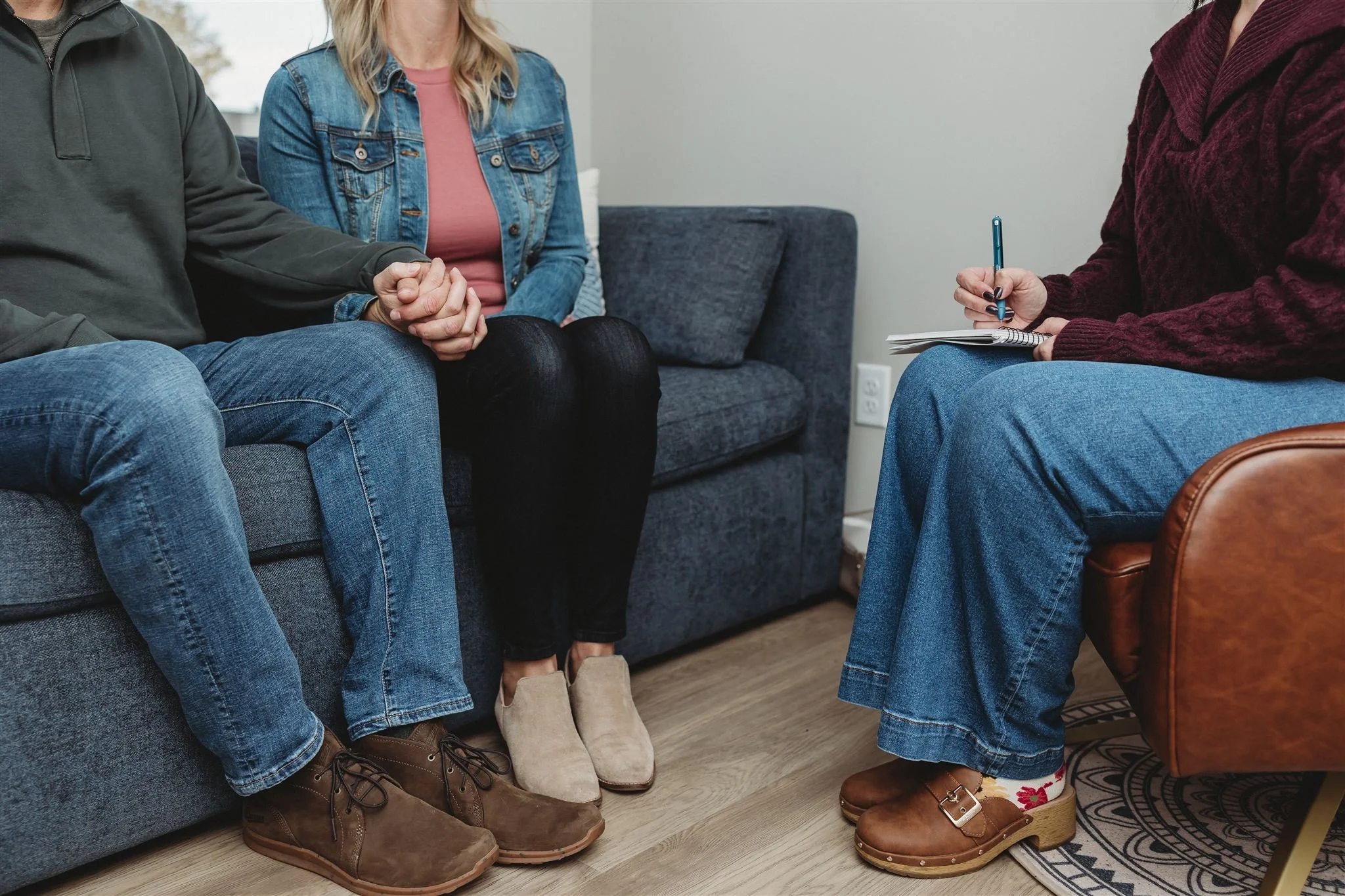 A couple sitting on a gray couch with a woman therapist taking notes during a counseling session.