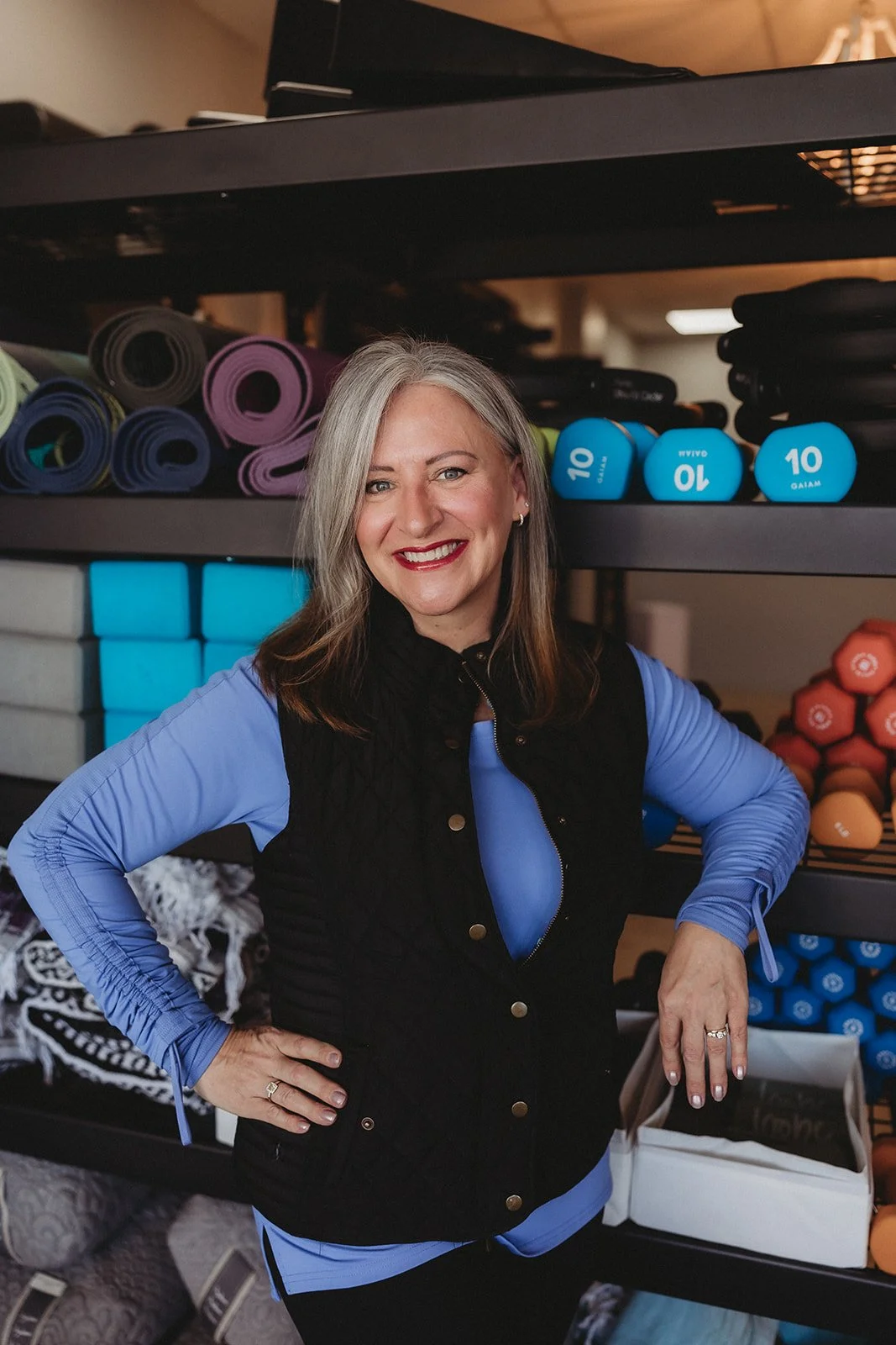 A woman with gray hair smiling, wearing a blue shirt and black vest, standing in front of shelves with colorful yoga mats and dumbbells in a gym or fitness store.