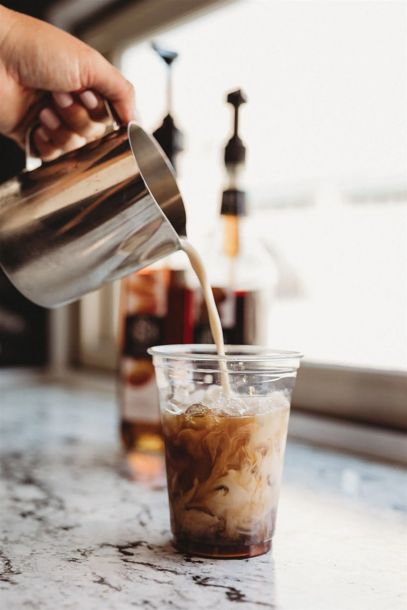 Person pouring milk into a glass of iced coffee or iced latte on a textured white countertop.