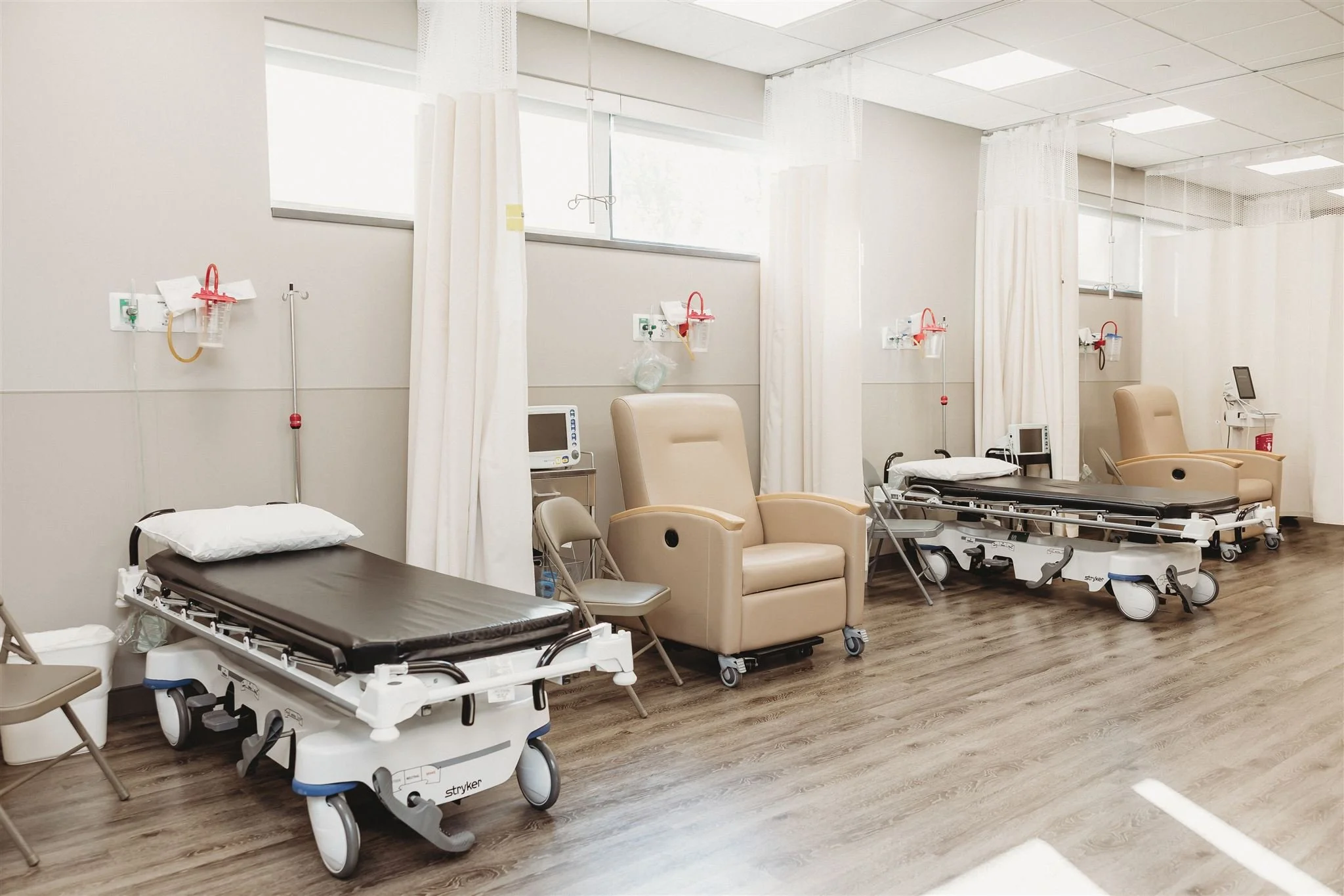 Empty hospital room with three beds and recliner chairs next to windows, medical equipment, and beige curtains.