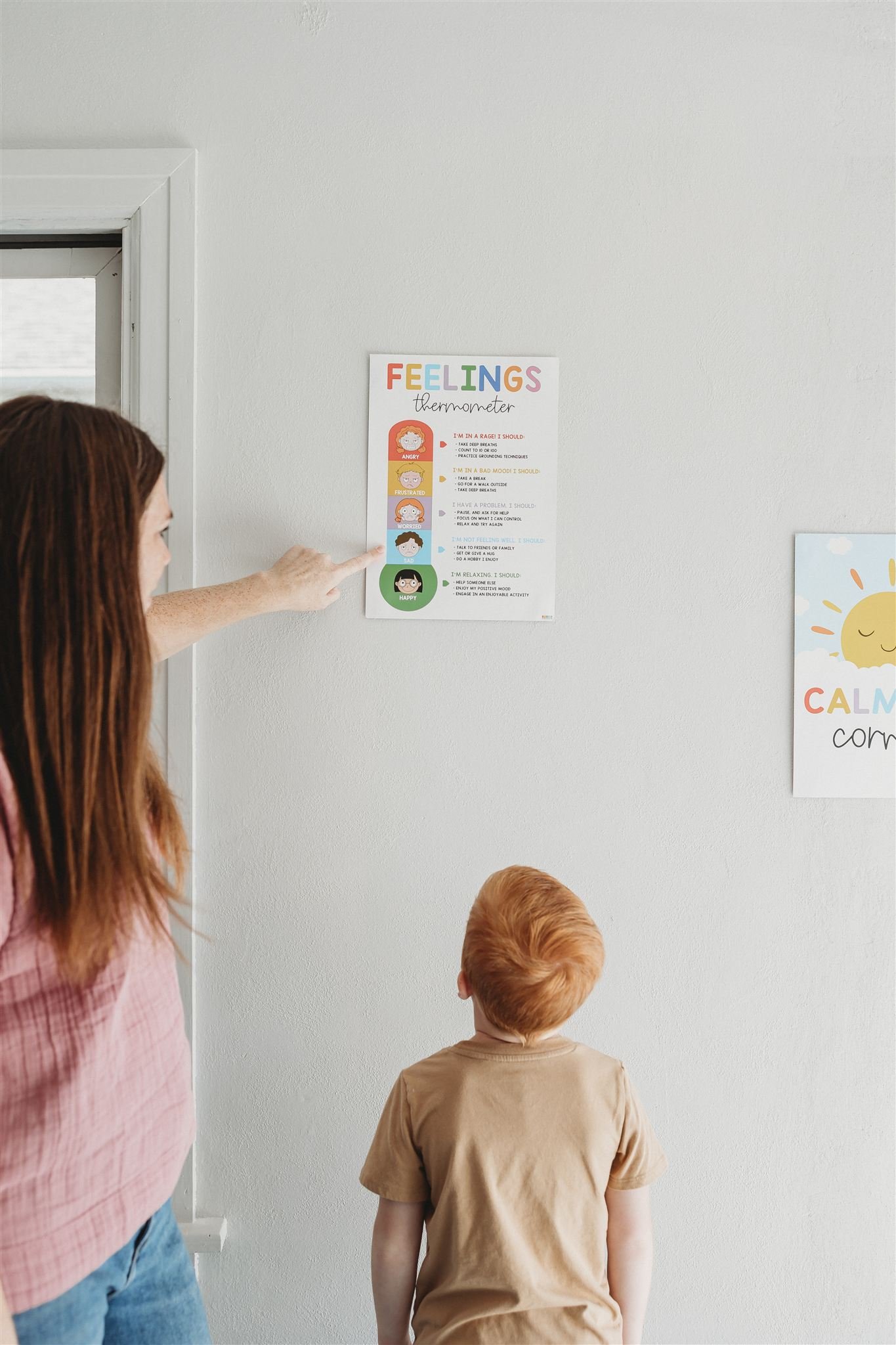 A woman pointing at a feelings thermometer chart on a white wall, with a young boy standing in front of her looking up at it. The chart features different feelings with corresponding colors and descriptions.