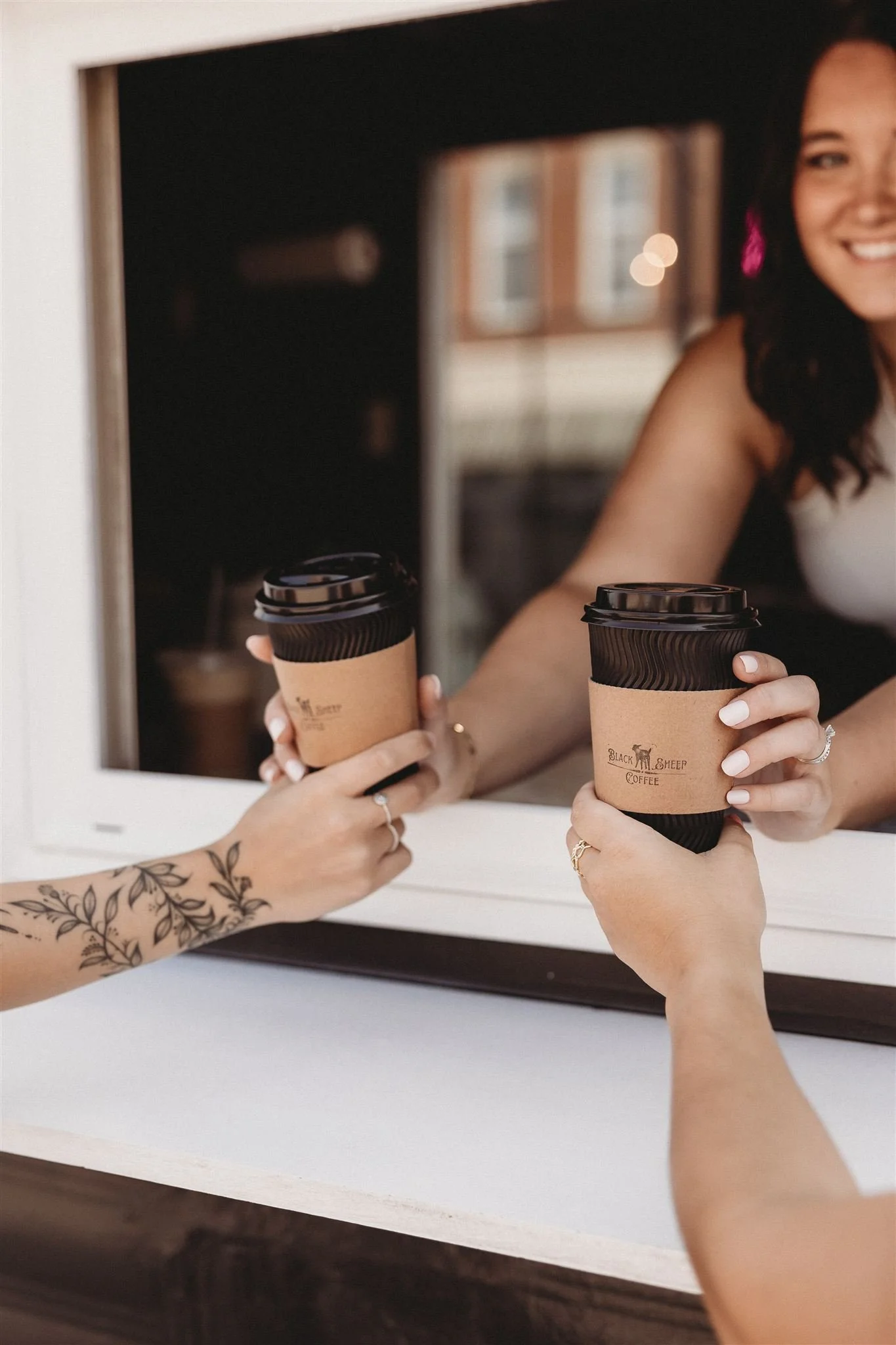 Two people exchanging coffee cups at a drive-thru window; one person has a floral tattoo on their arm, and both are wearing rings.