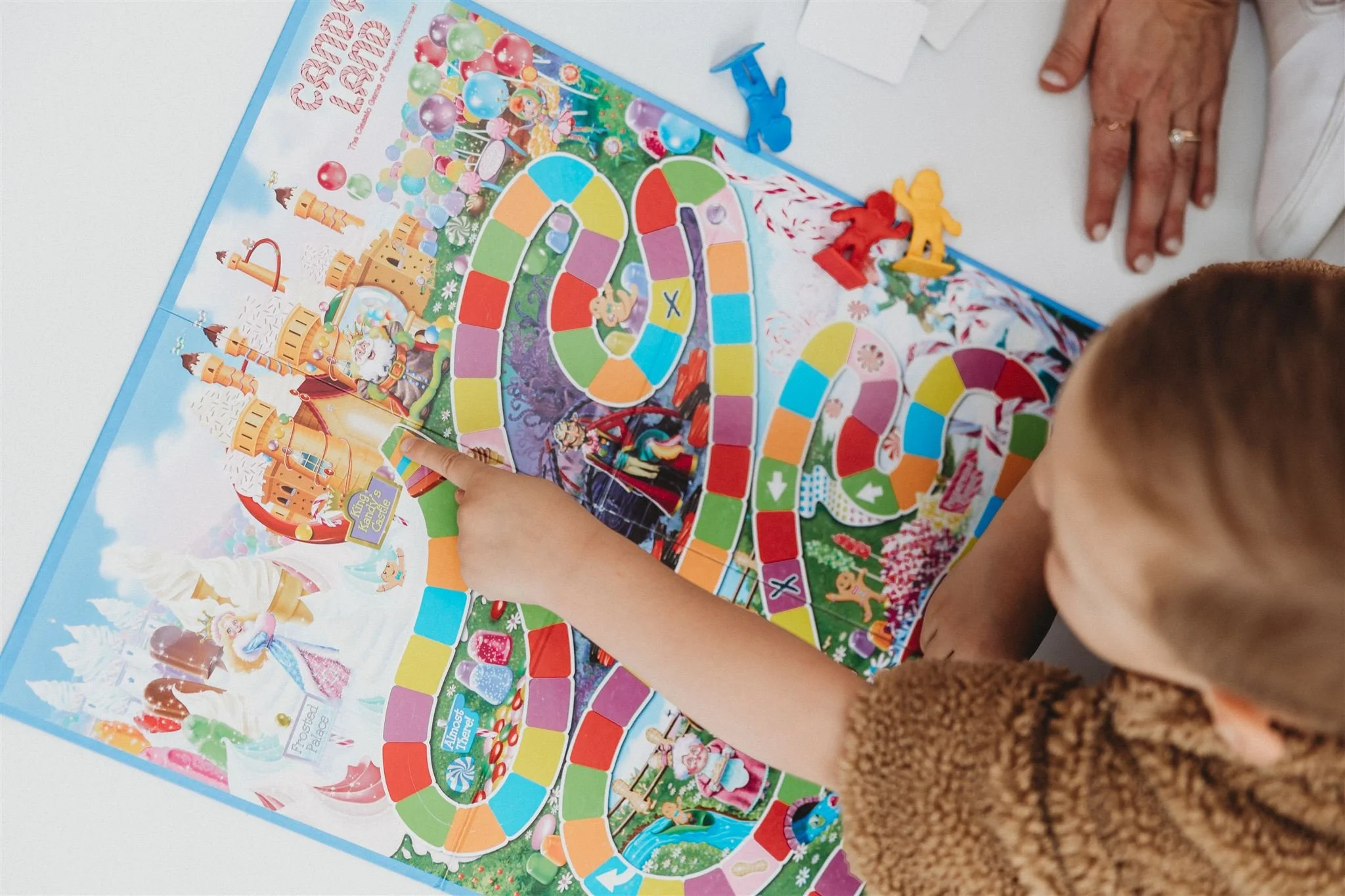 A child points at a colorful Candy Land board game spread out on a table, with bright winding paths, candy-themed illustrations, and game pieces nearby. An adult’s hand rests beside the board.