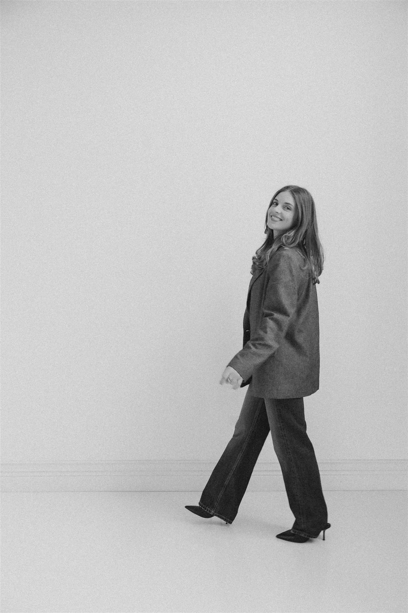 Black and white image of a woman walking with a smile, wearing a blazer and flared jeans, against a plain background.