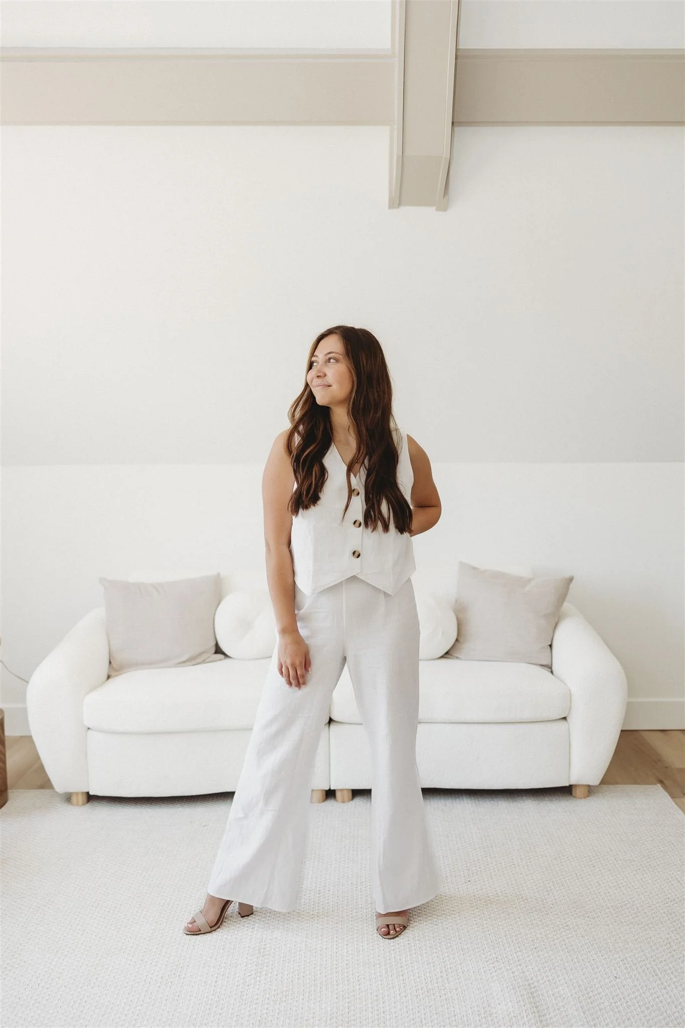 A woman standing in front of a white sofa with pillows in a bright, minimalist room, wearing a sleeveless white top and white pants.