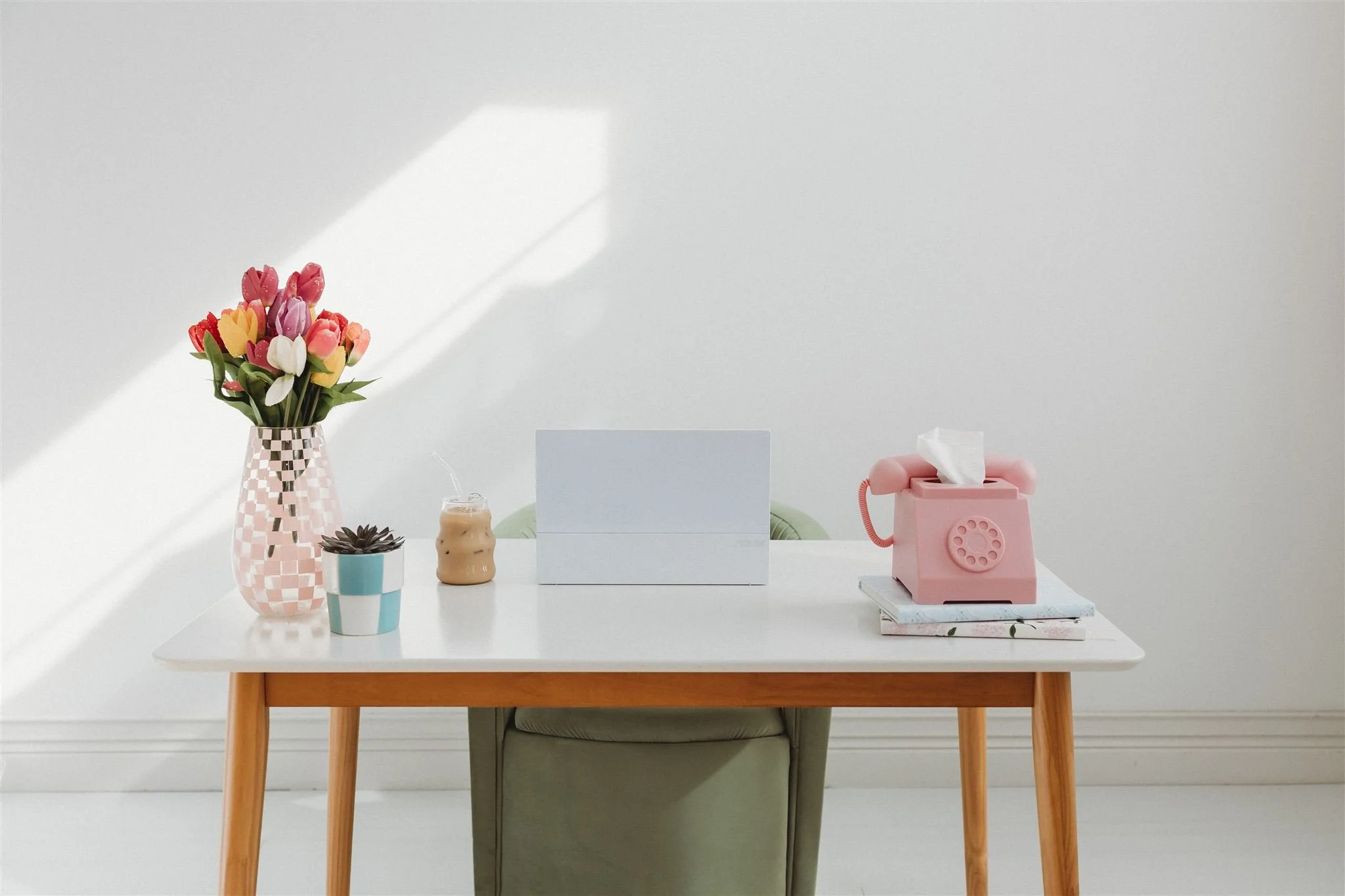 A minimal styled desk with a white laptop, tulips, iced coffee, and a pink retro telephone tissue box, bathed in natural light.