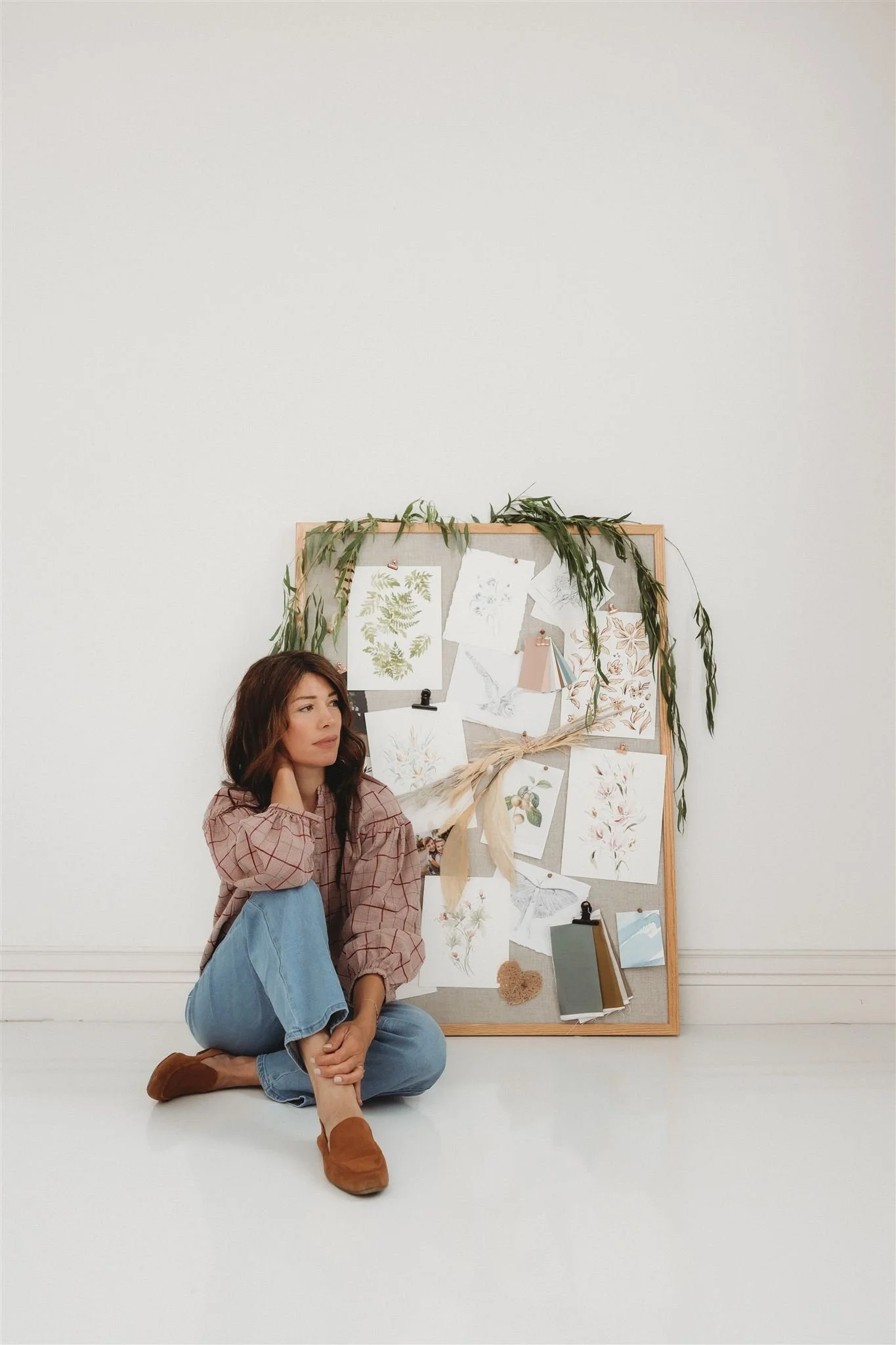 A woman sitting on the floor beside a large corkboard decorated with botanical illustrations, drawings, and notes, some pinned with clips and a tied ribbon, with green leafy garland draped over the top.