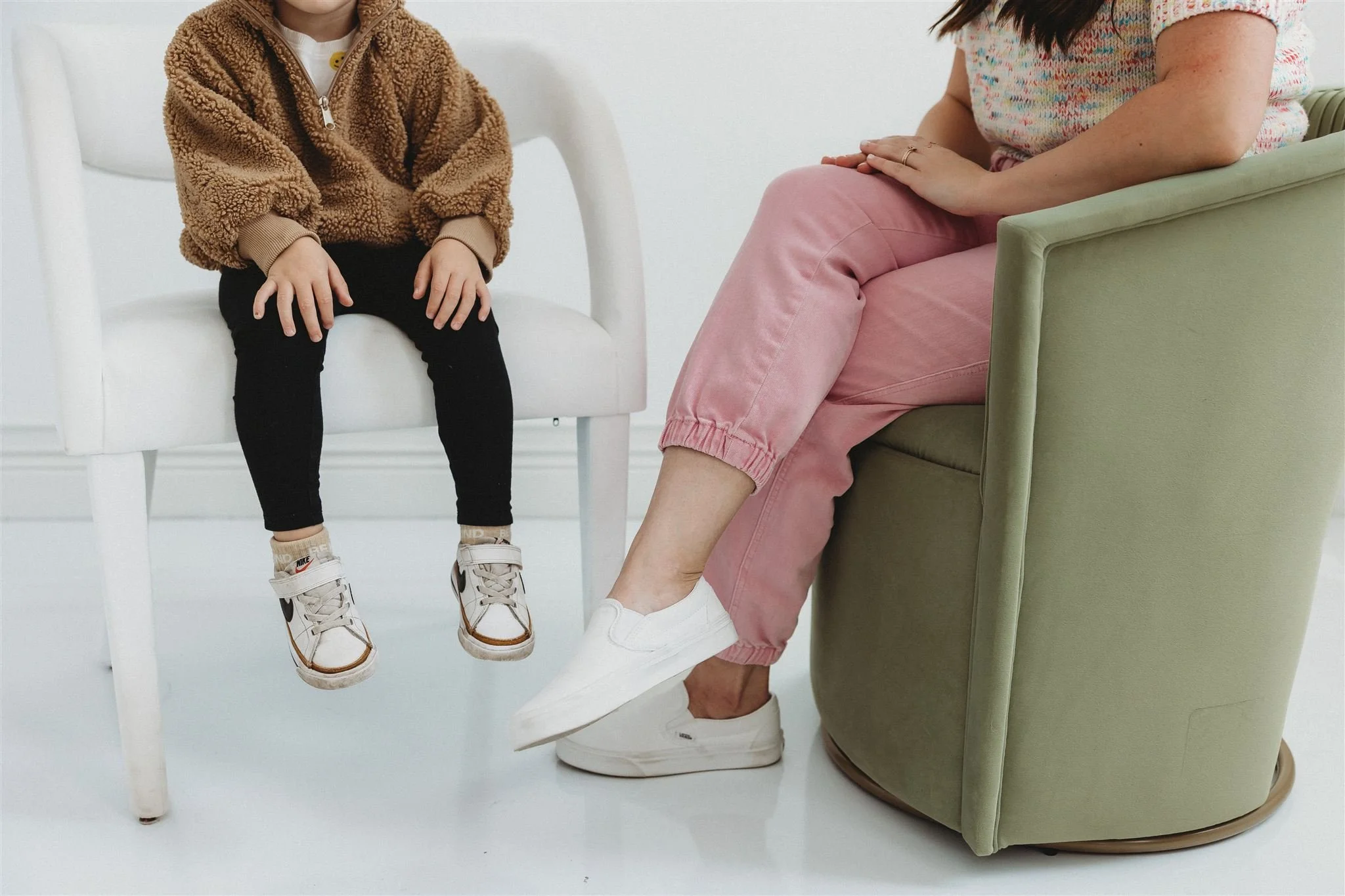 Alt-text: A child in a brown fleece jacket and Nike sneakers sits in a white chair across from an adult in pink joggers and white slip-on sneakers seated in a sage green chair, both with faces out of frame.