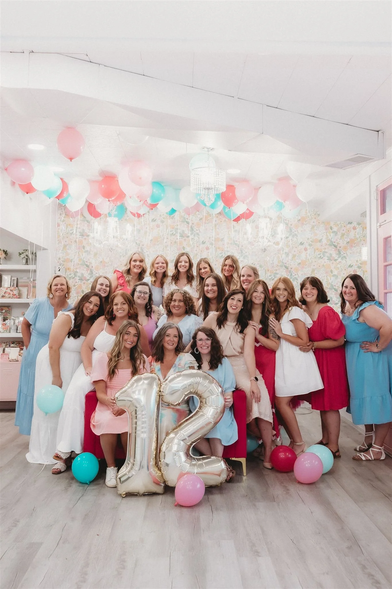 Group of women celebrating birthday with balloons, including large silver balloon numbers 1 and 2, in a decorated room with pastel balloons and floral wallpaper.