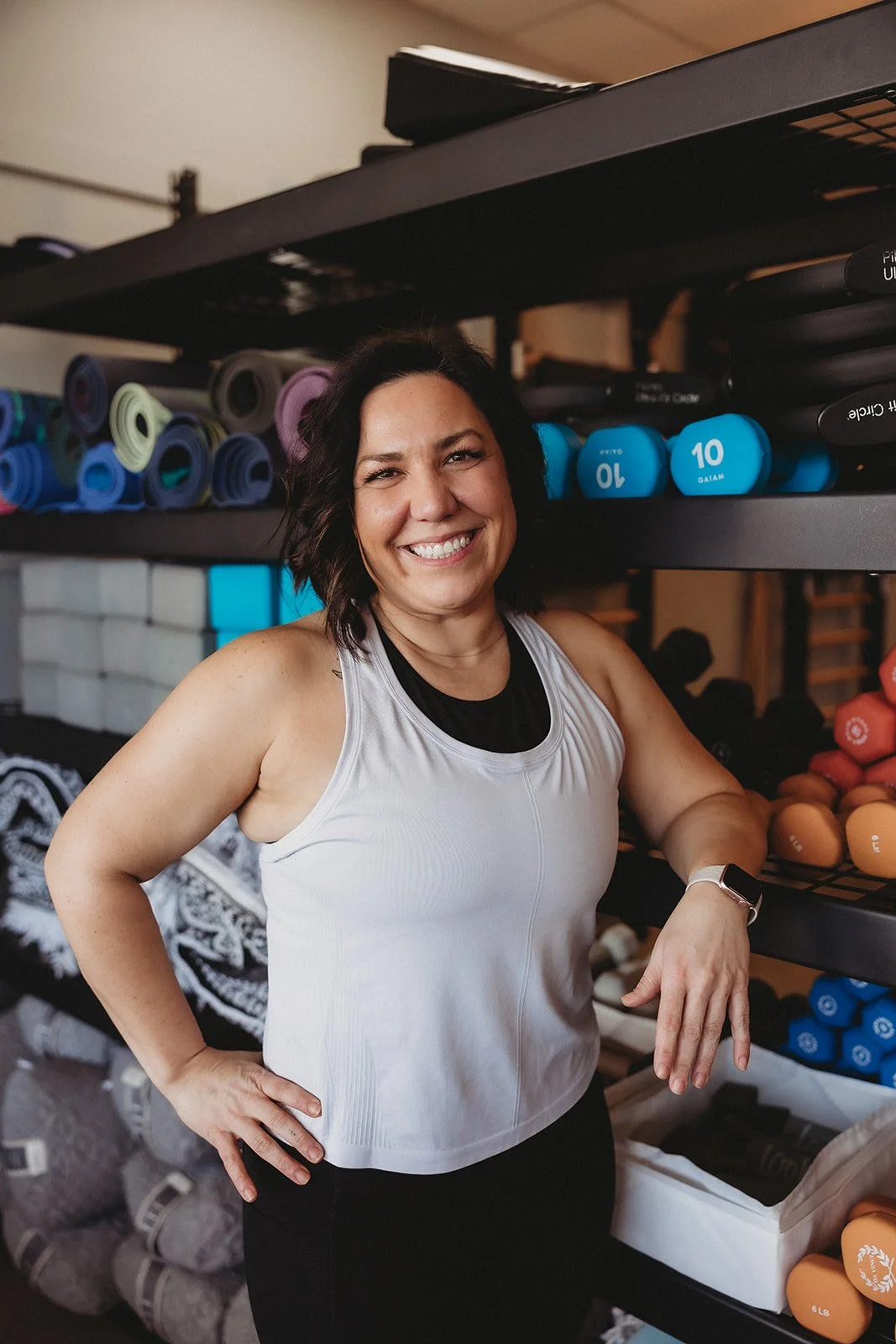 A woman with dark hair and a white workout top standing in a gym with colorful dumbbells and workout equipment on shelves behind her, smiling at the camera.