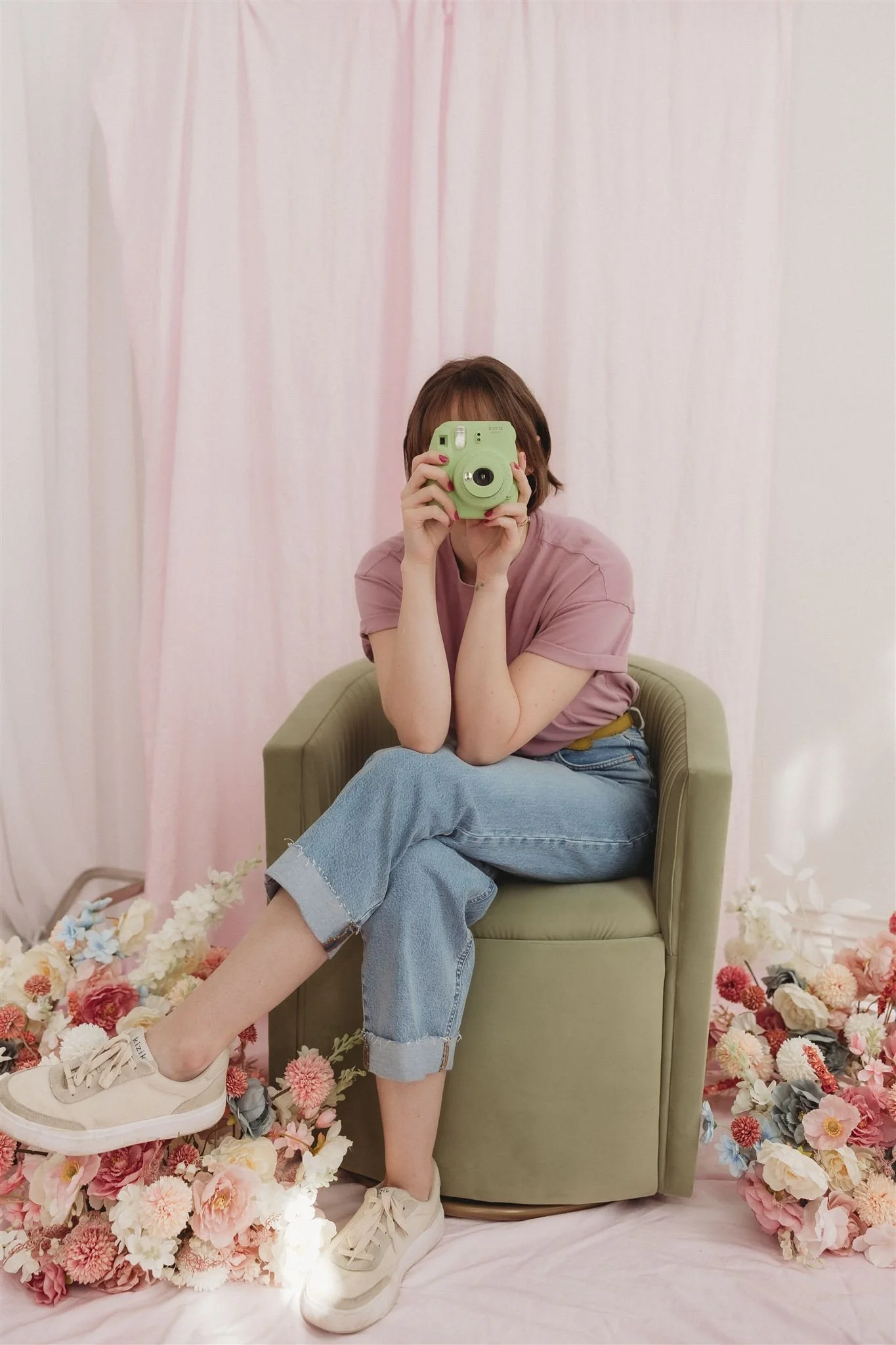 Person taking a mirror selfie, sitting on a green armchair with pink curtains and floral decorations in the background.