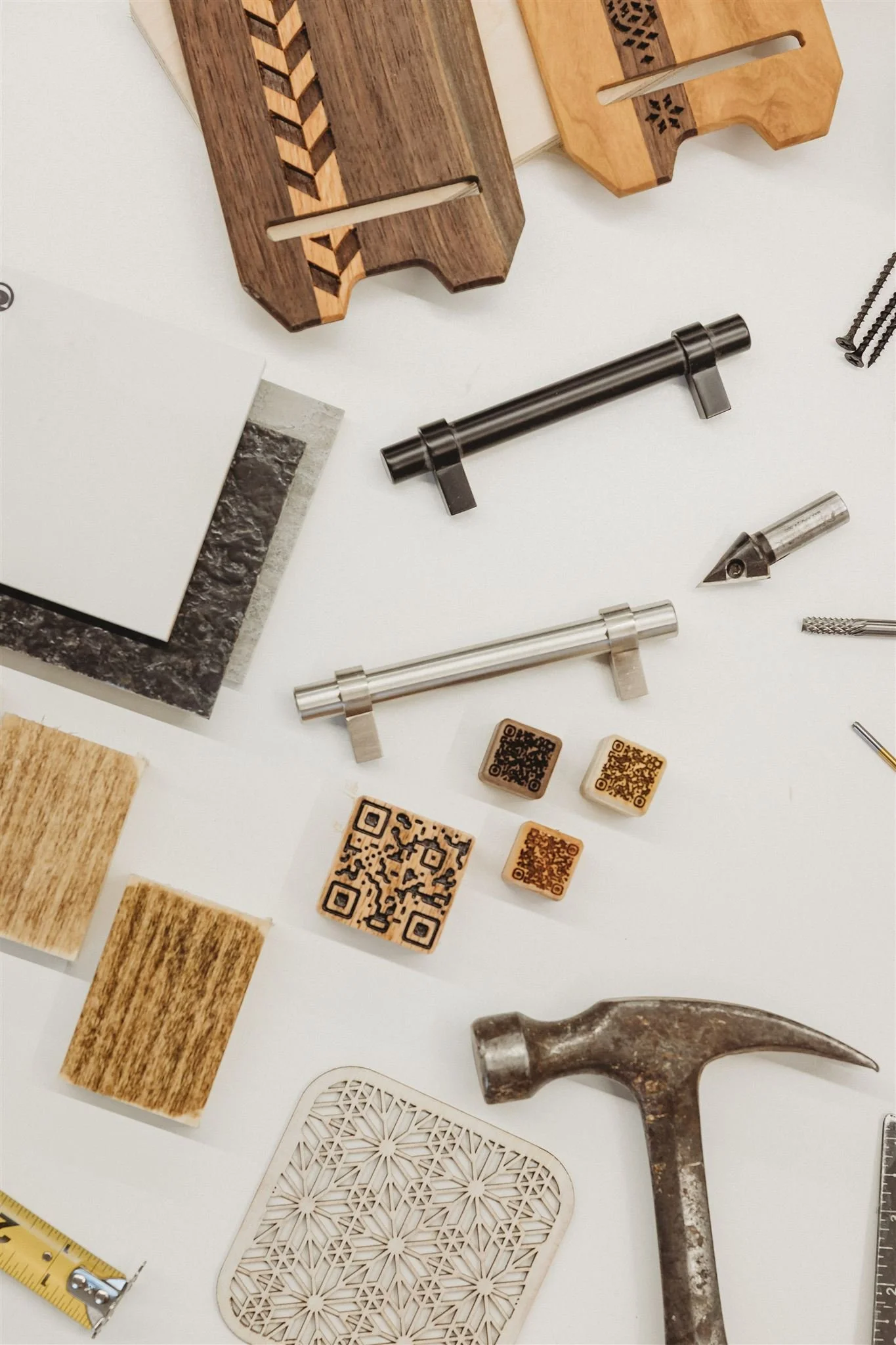 A flat lay of woodworking tools and materials, including wooden slats, metal clamps, printing blocks, a hammer, screws, a ruler, and QR code stamps on a white surface.