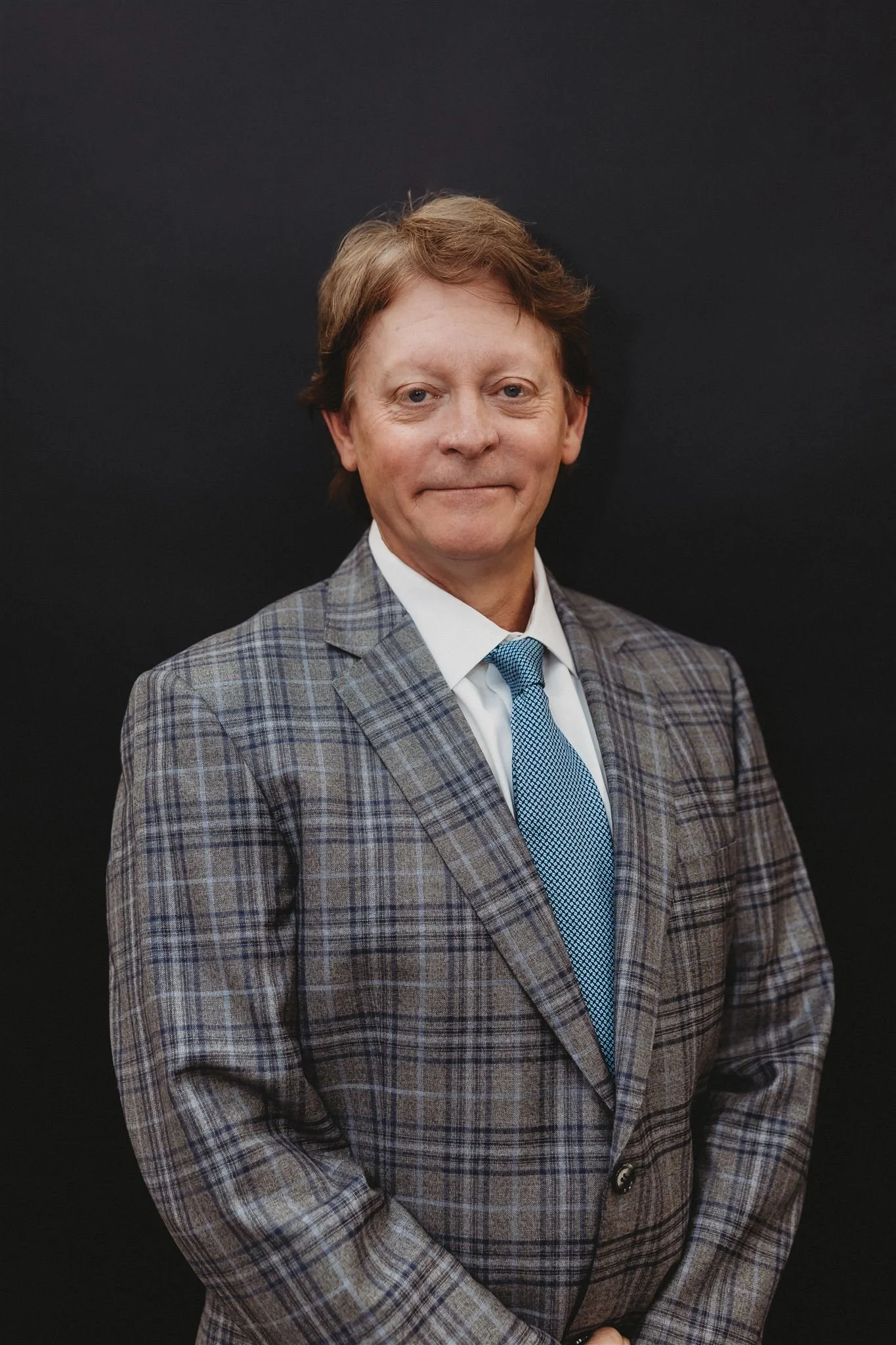 A middle-aged man with light skin, wearing a gray plaid suit, white shirt, and blue tie, standing against a plain black background.