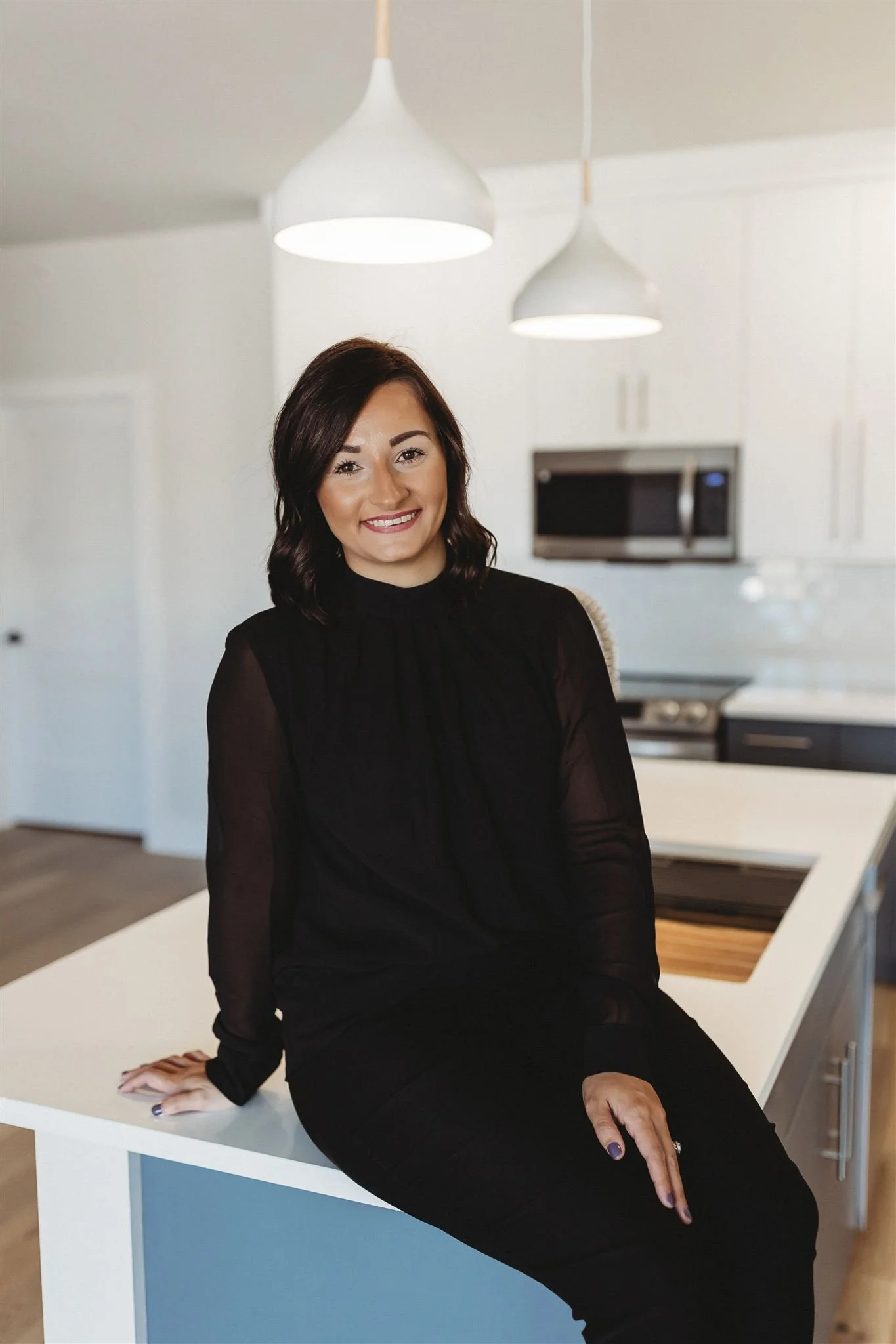 A woman with dark hair wearing a black long-sleeve top, sitting on a kitchen island in a modern white kitchen with hanging pendant lights and stainless steel appliances.