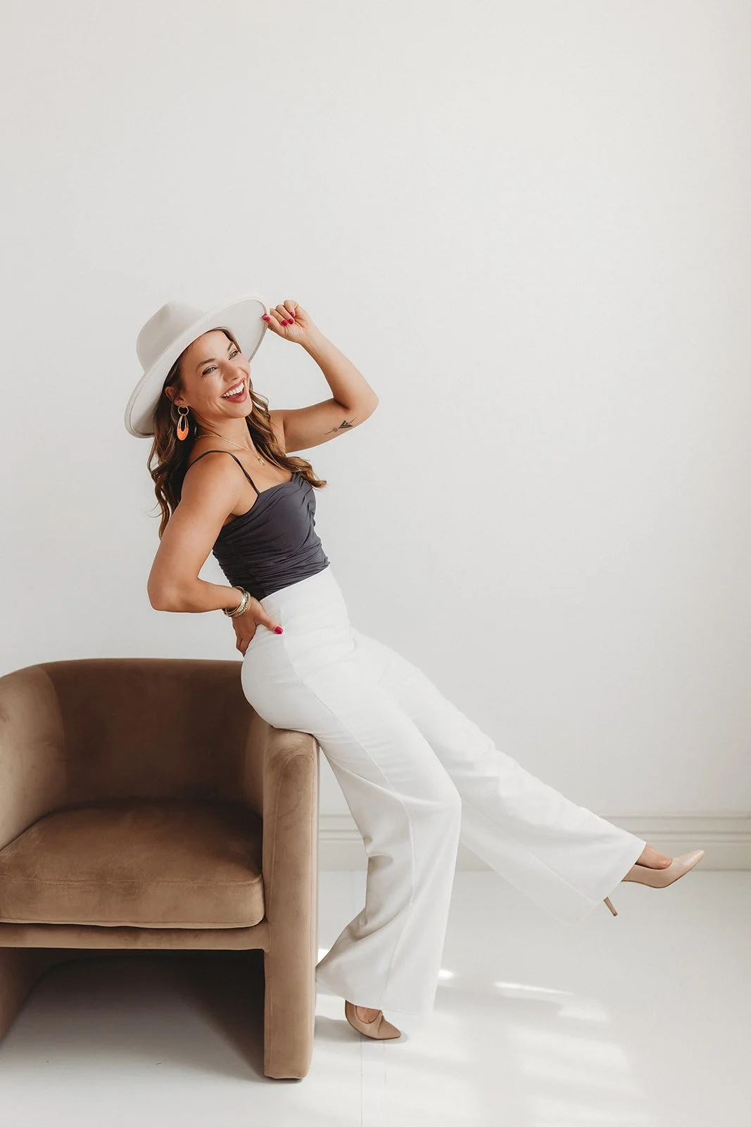 Woman in a black top and white wide-leg pants, wearing a white hat, standing next to a beige armchair in a minimalist room, smiling and posing playfully.