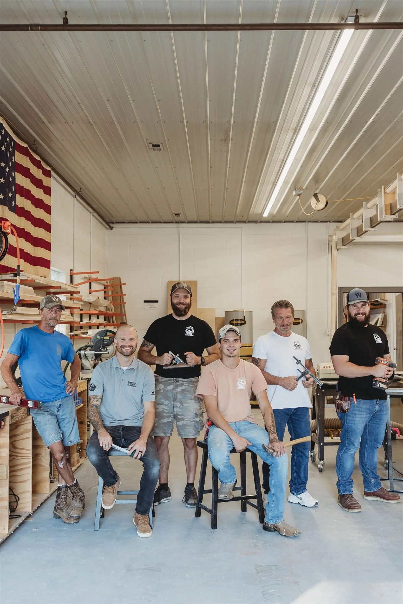 Six men inside a woodworking shop, some holding tools, with woodwork supplies and an American flag on the wall.