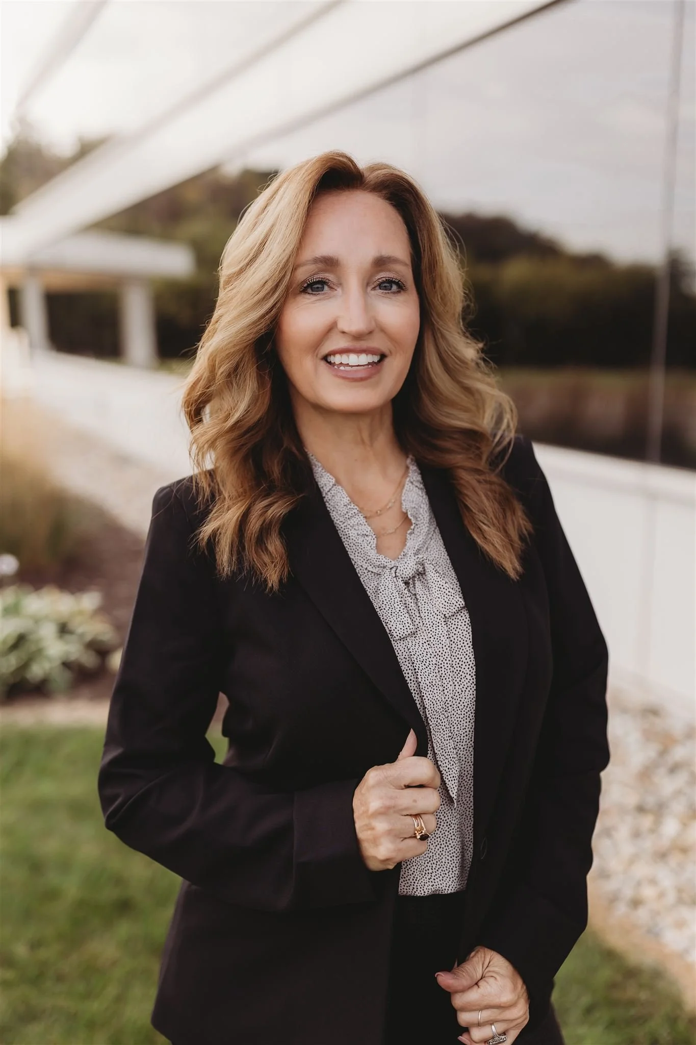 A woman with long, wavy blond hair smiling outdoors in front of a modern building with reflective glass windows, wearing a black blazer over a patterned blouse.