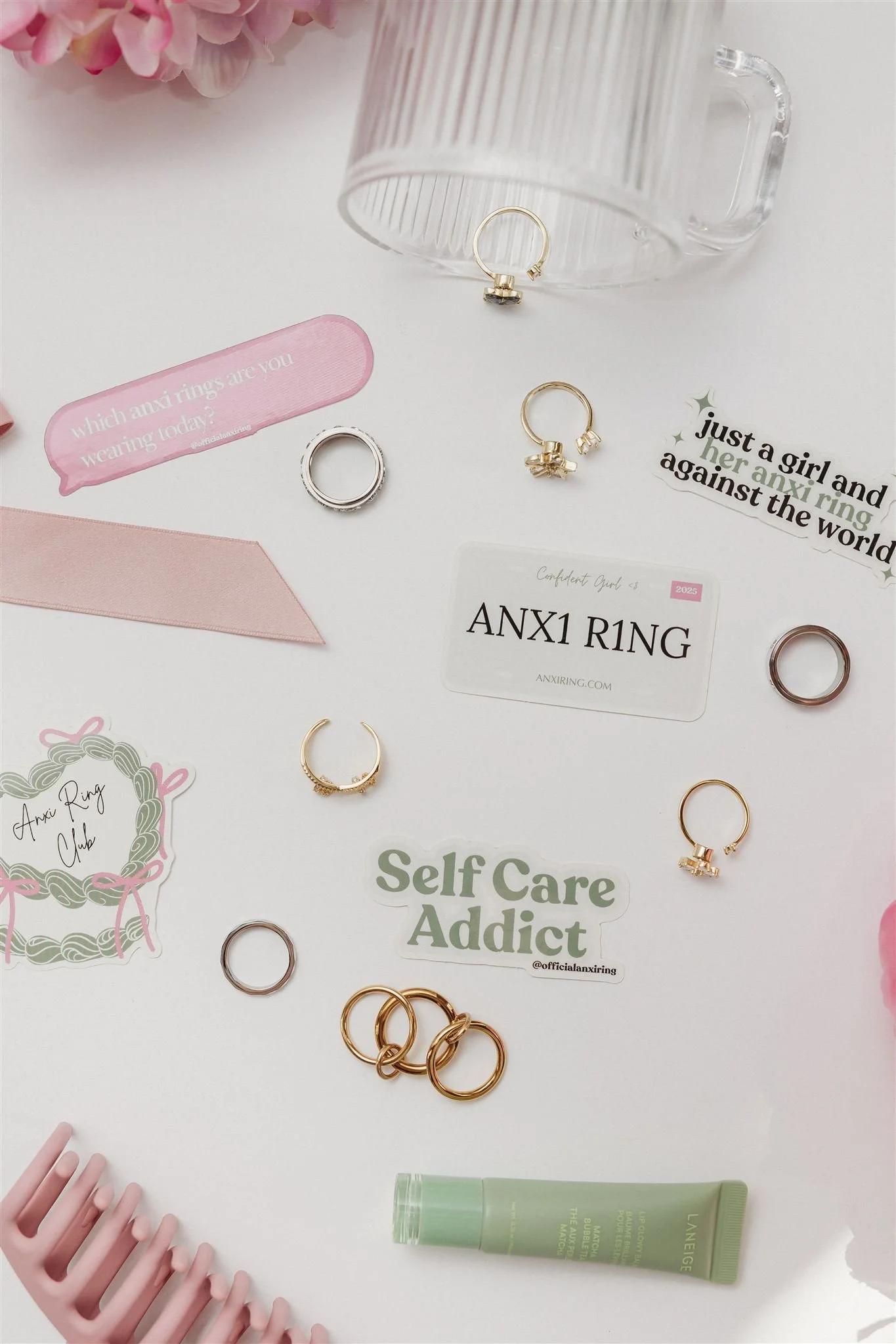 A collection of jewelry rings, a pink hair comb, skincare lip balm, and decorative stickers on a white surface. There is also a glass mug and various small accessories.