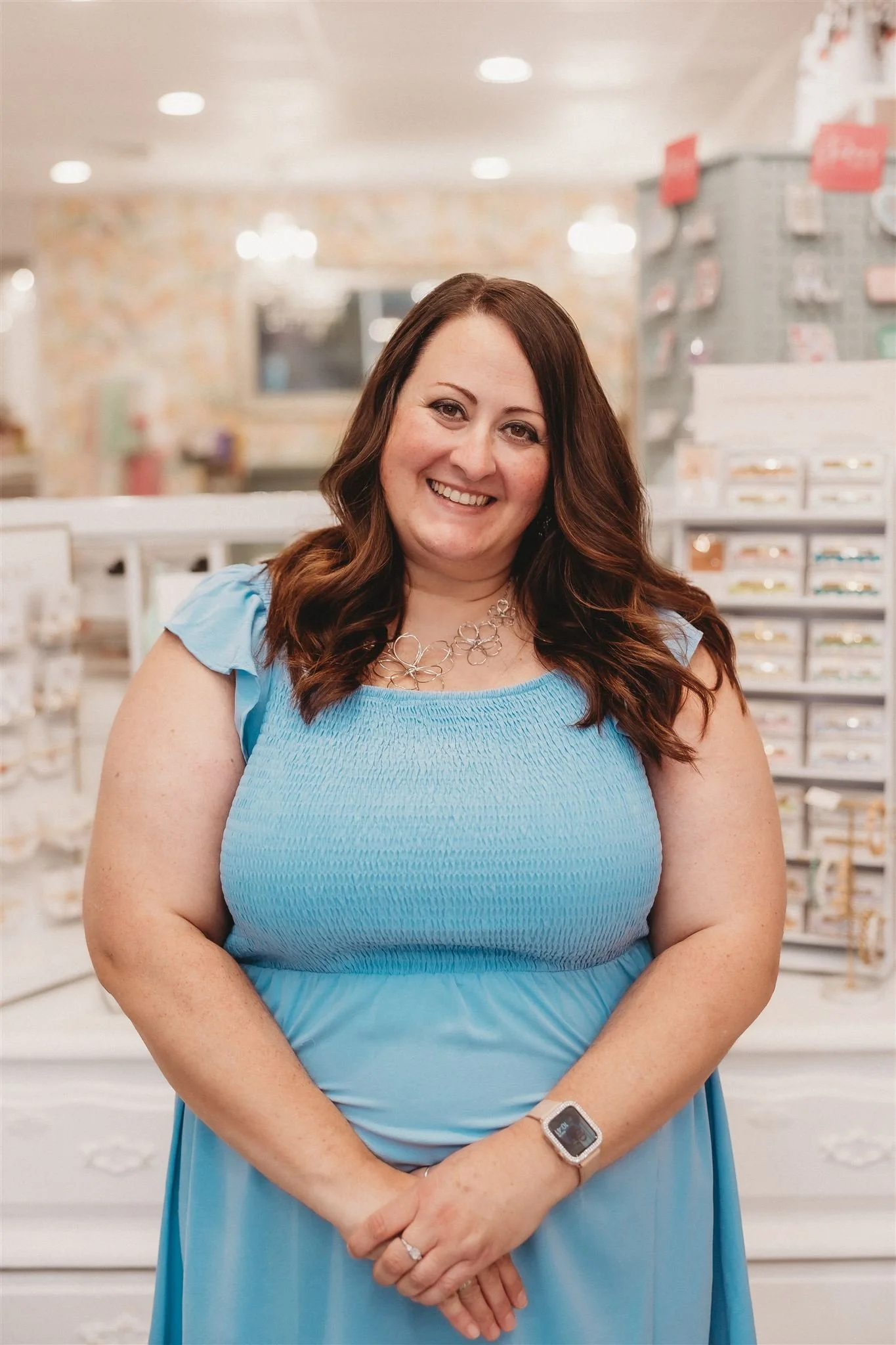 A woman with brown wavy hair wearing a blue dress, a necklace, and a smartwatch, smiling in a store with jewelry displays in the background.