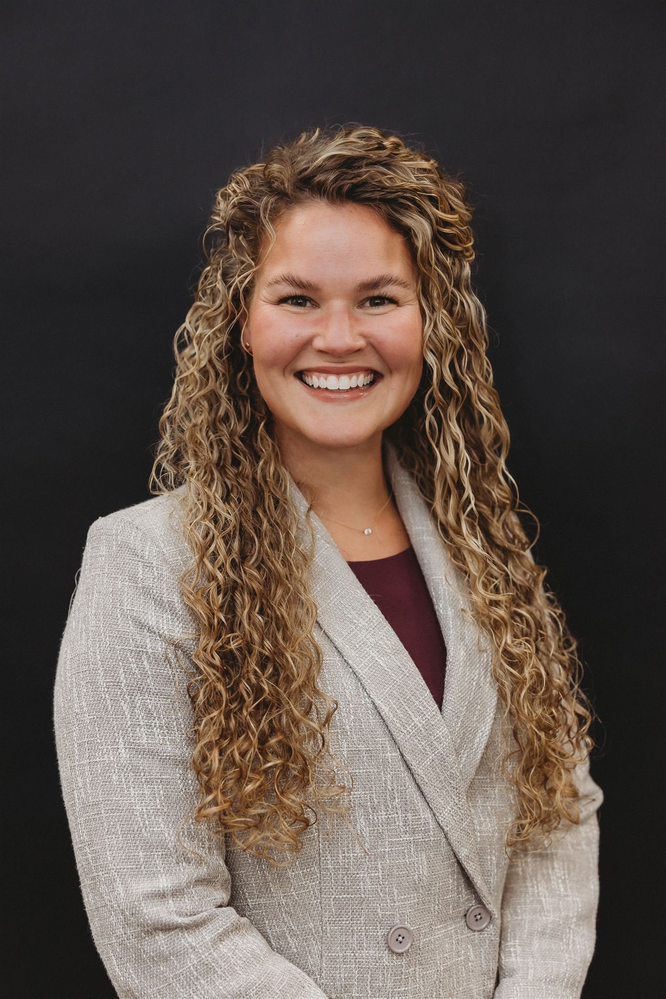 Professional woman with long curly blonde hair, wearing a beige blazer over a burgundy top, smiling against a dark background.
