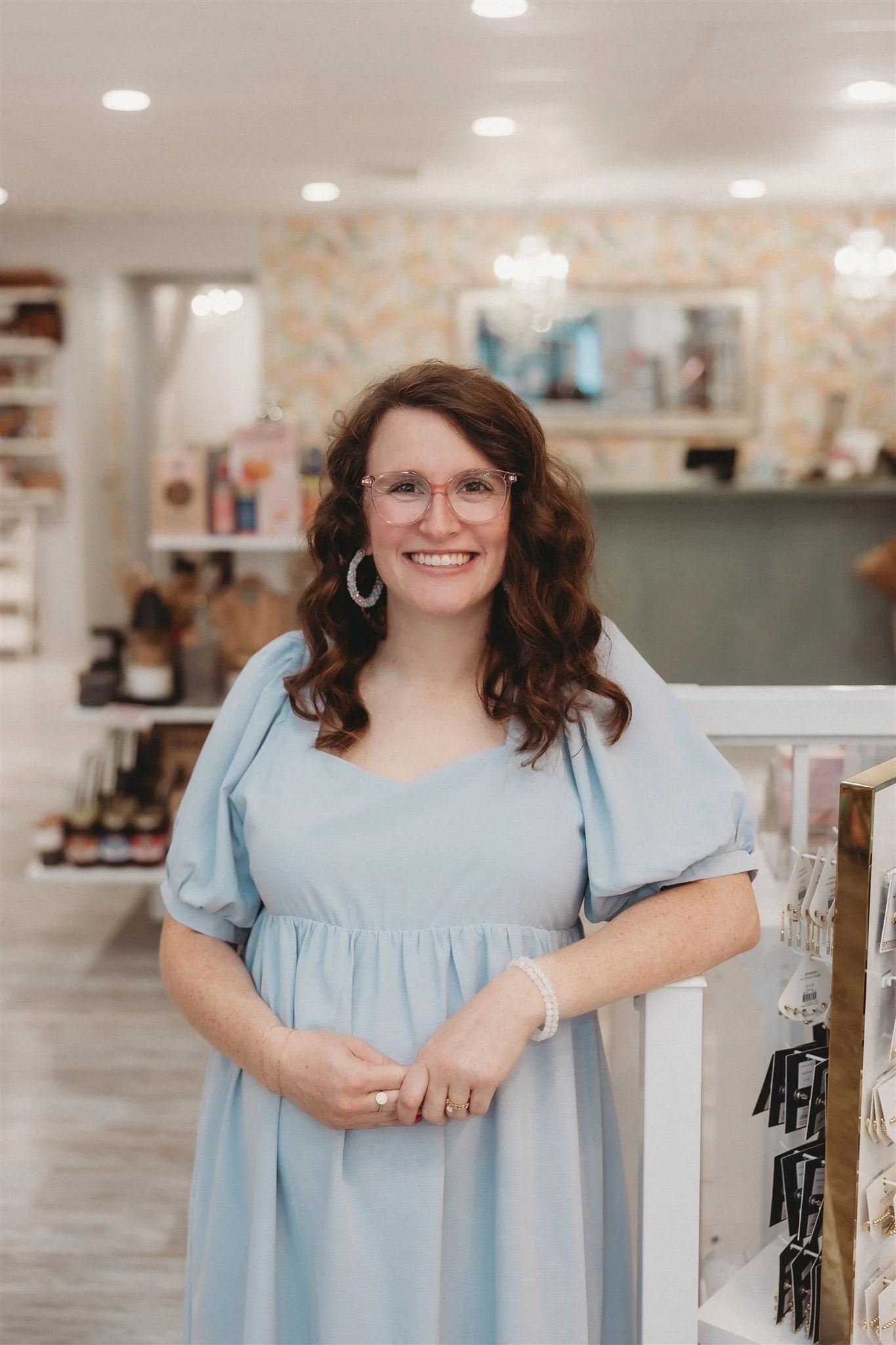 A smiling woman with brown curly hair, pink glasses, and hoop earrings in a light blue dress standing inside a store.
