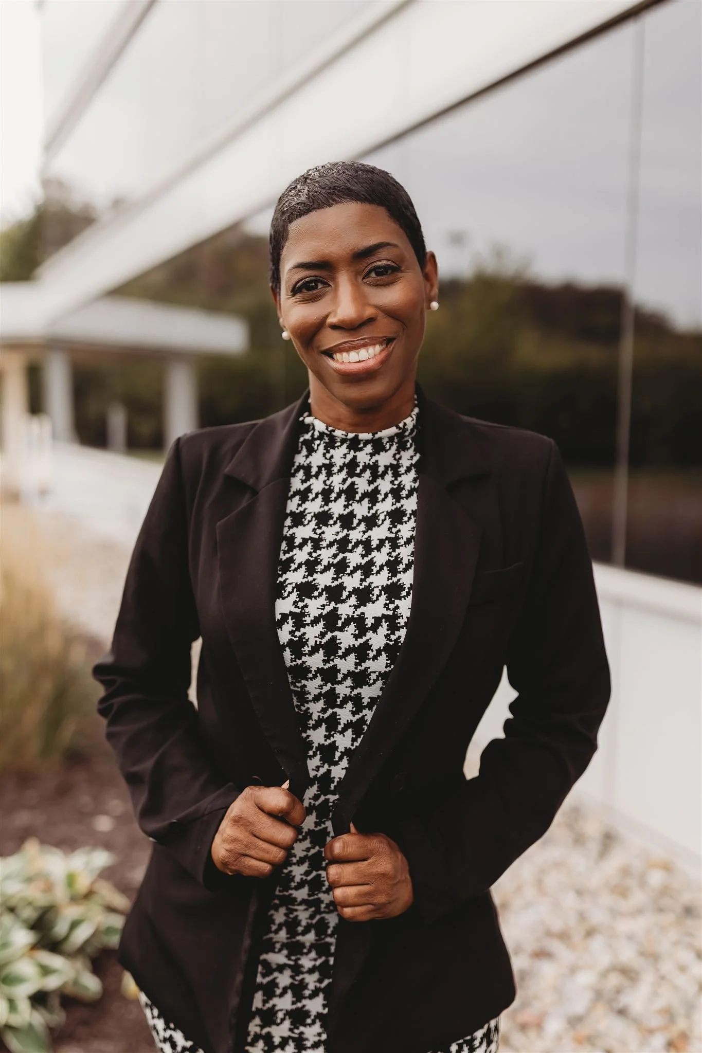 A woman with short black hair and a bright smile, wearing a black blazer over a houndstooth-patterned top, standing outdoors near a modern glass building with blurred natural background.