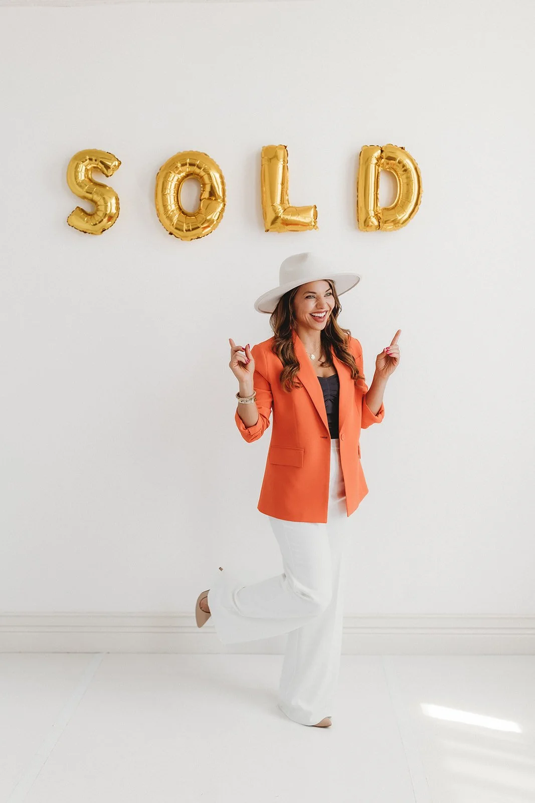 Woman in orange blazer and white pants standing on one leg in front of a white wall with gold balloon letters spelling "SOLD", wearing a white hat and smiling.