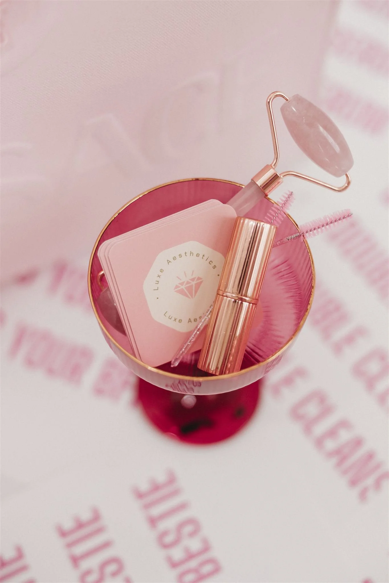 Pink cosmetic display with a jade roller, pink mascara, a container of pink cotton swabs, and a stack of pink cards, set on a pink surface.