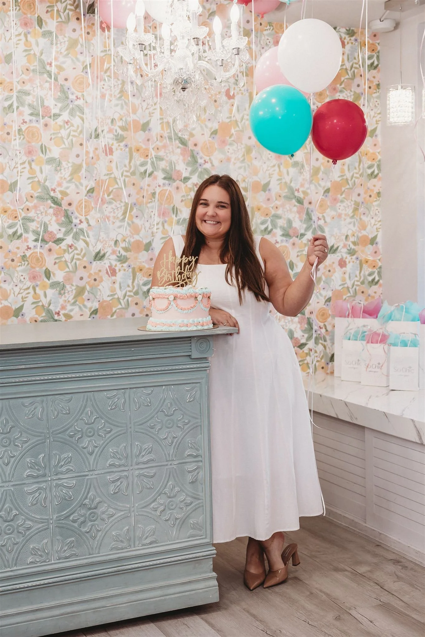 A woman in a white dress celebrating her birthday with a small decorated cake, balloons, and gift bags in the background, standing by a vintage turquoise counter in a floral decorated room.