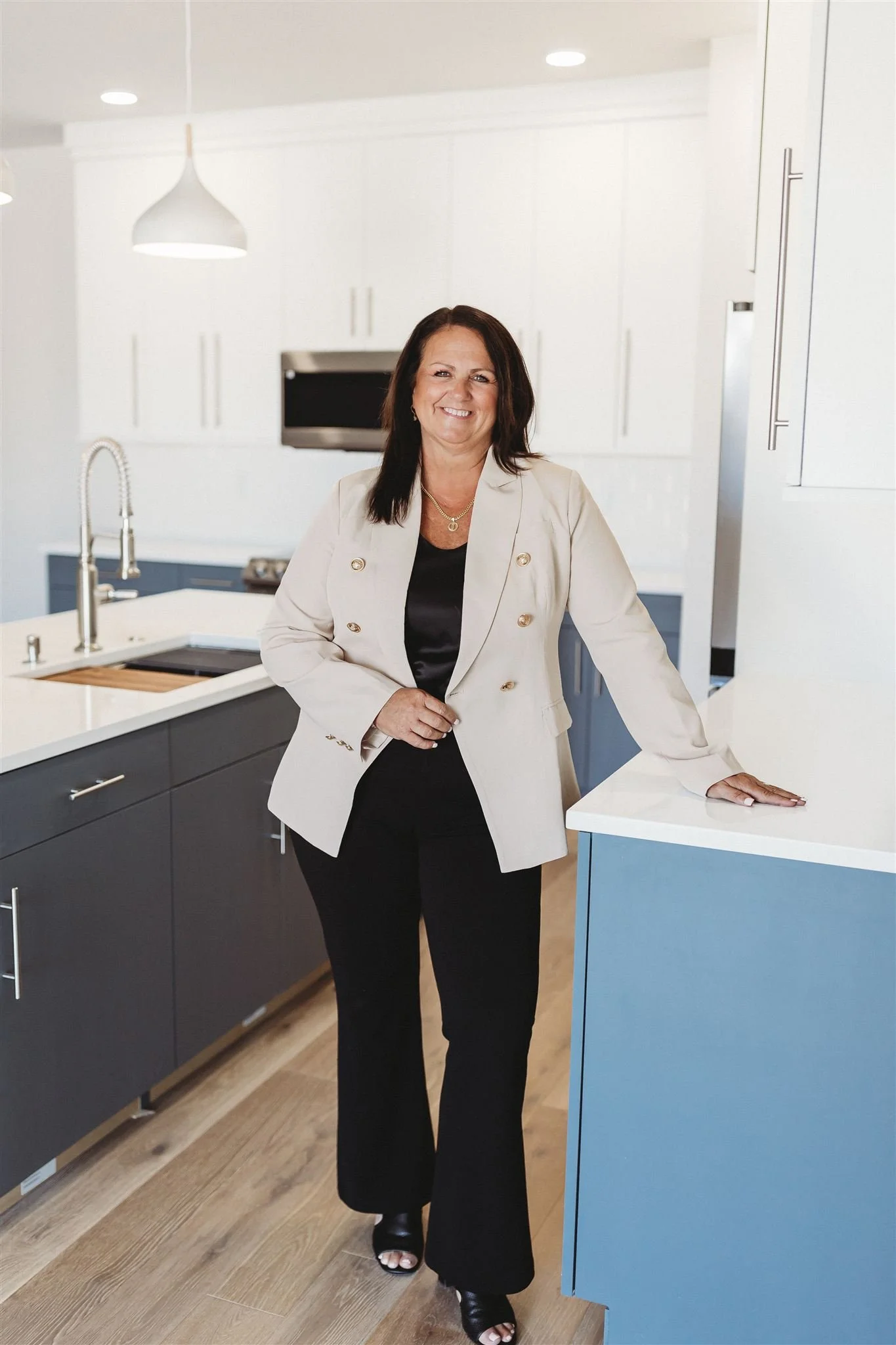A woman standing in a modern kitchen with white cabinets and a gray island, smiling and wearing a white blazer, black top, and black pants.