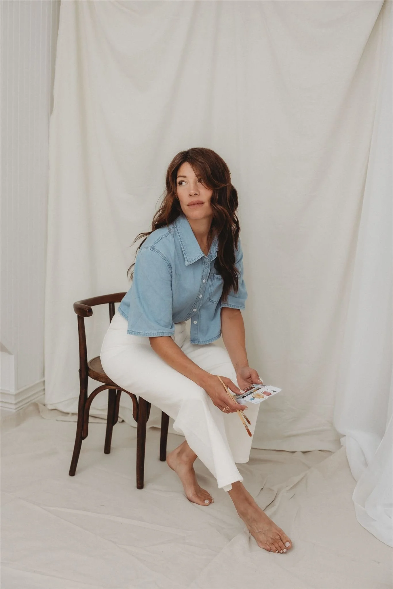 A woman with brown wavy hair in a blue denim shirt and white pants is sitting barefoot on a wooden chair, holding a palette and brush, with a neutral expression, in front of cream-colored curtains and a white room.