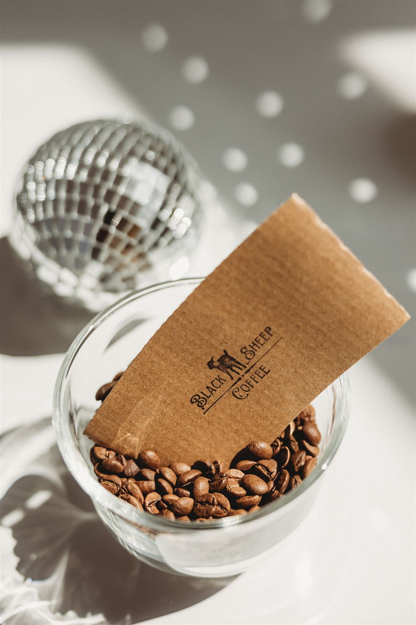 A glass bowl filled with roasted coffee beans, with a brown paper tag reading 'Black Sheep Coffee' inserted into the beans, a small disco ball reflecting light, and a dotted shadow pattern on the white surface.