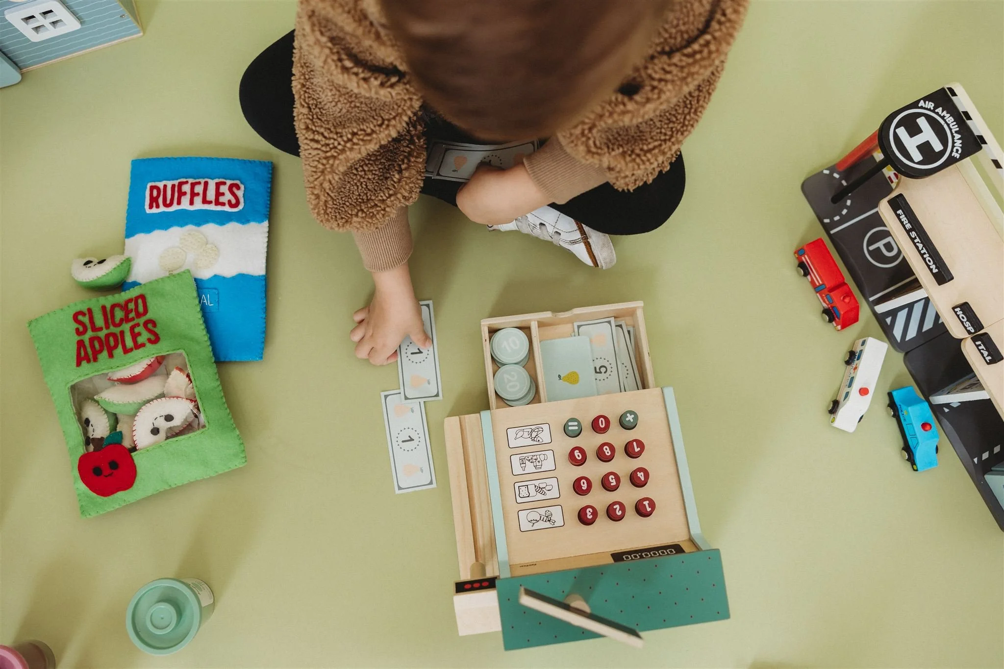 A child sits on the floor playing with a toy cash register, arranging play money and coins. Nearby are felt play food items labeled ‘ruffles’ and ‘sliced apples,’ along with small toy vehicles and a play mat.