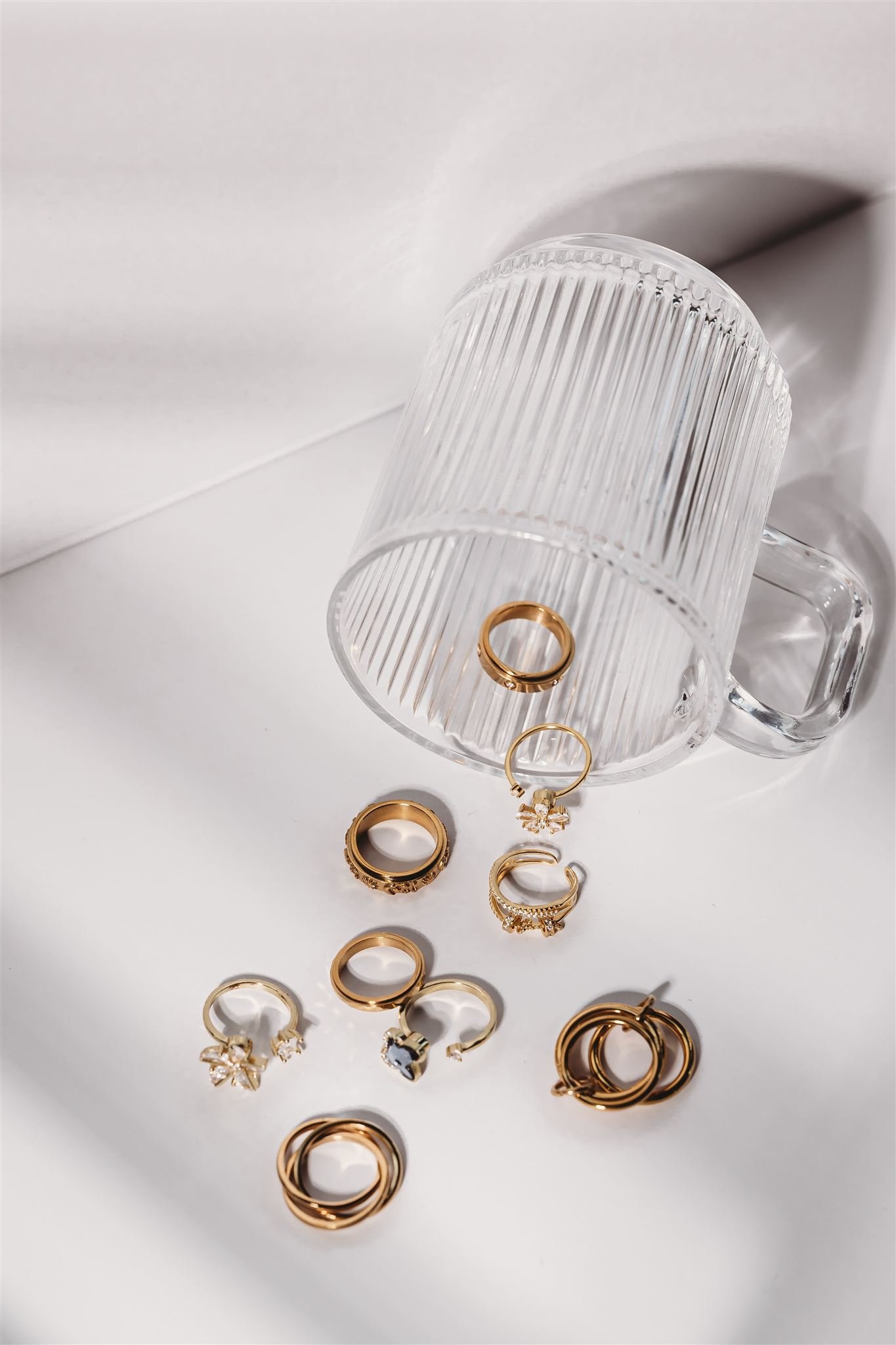 Gold and silver rings scattered on a white surface, with some of them partially inside a clear ribbed glass mug.