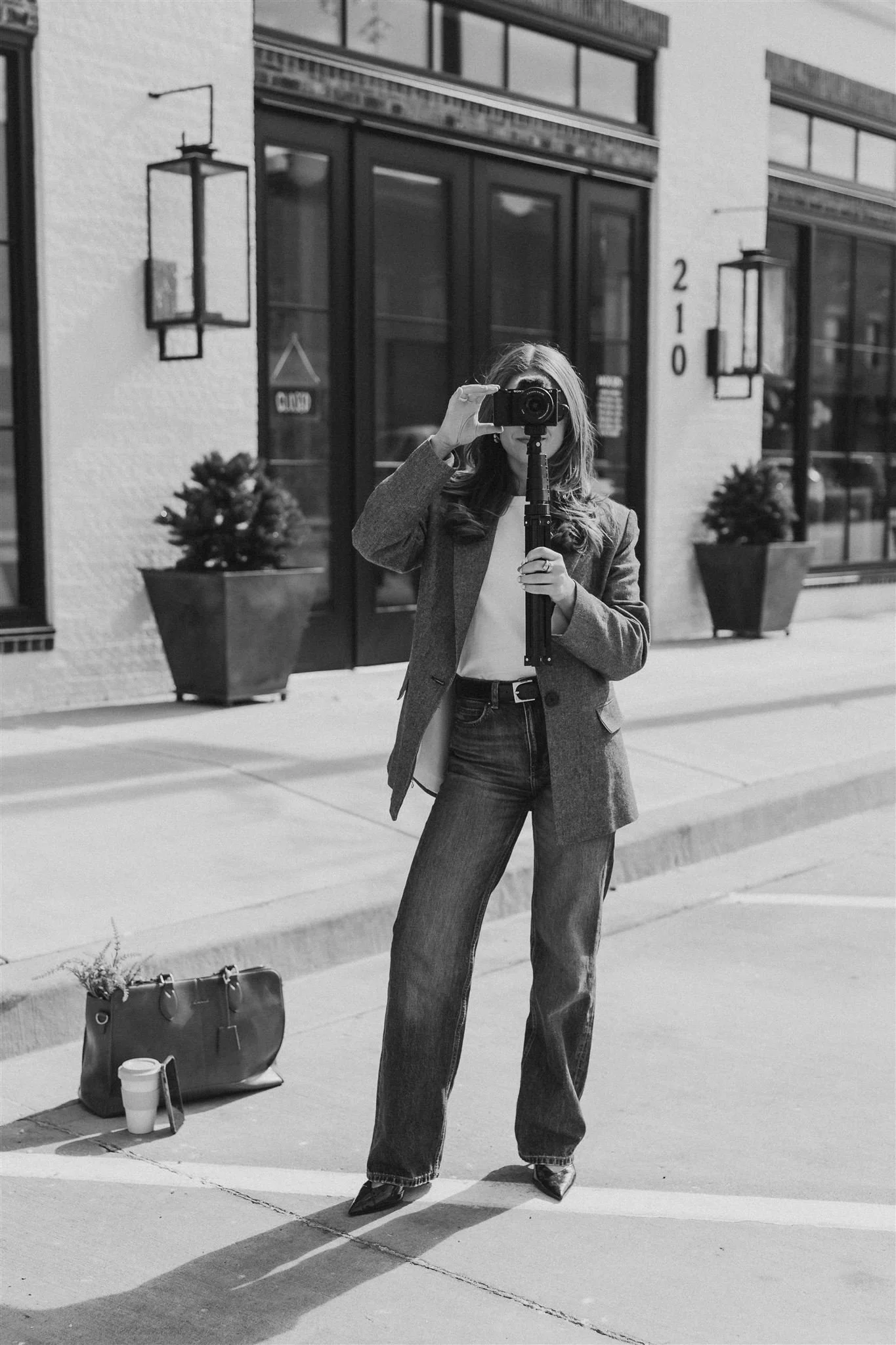 A woman taking a photo with a camera on a tripod on a city sidewalk in black and white. She is wearing jeans, a blazer, and pointed shoes. There are potted plants, a coffee cup, and a handbag on the ground nearby, with storefronts and street lamps in the background.