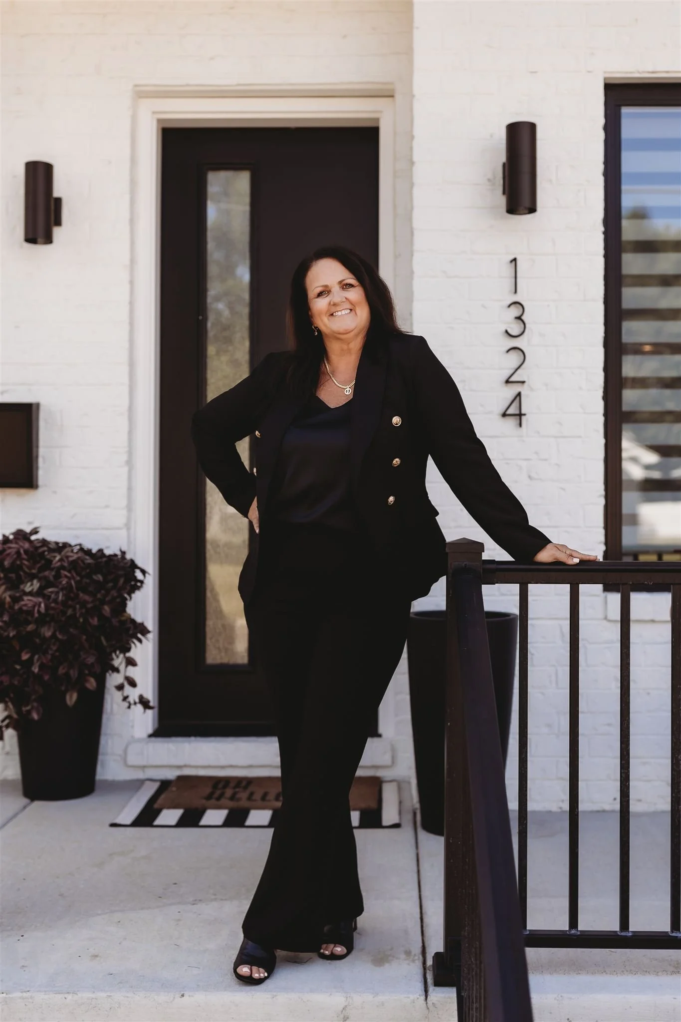 A woman standing on a front porch in front of a modern house with white brick walls, a black door, and the house number 1324. She is wearing a black outfit and smiling.