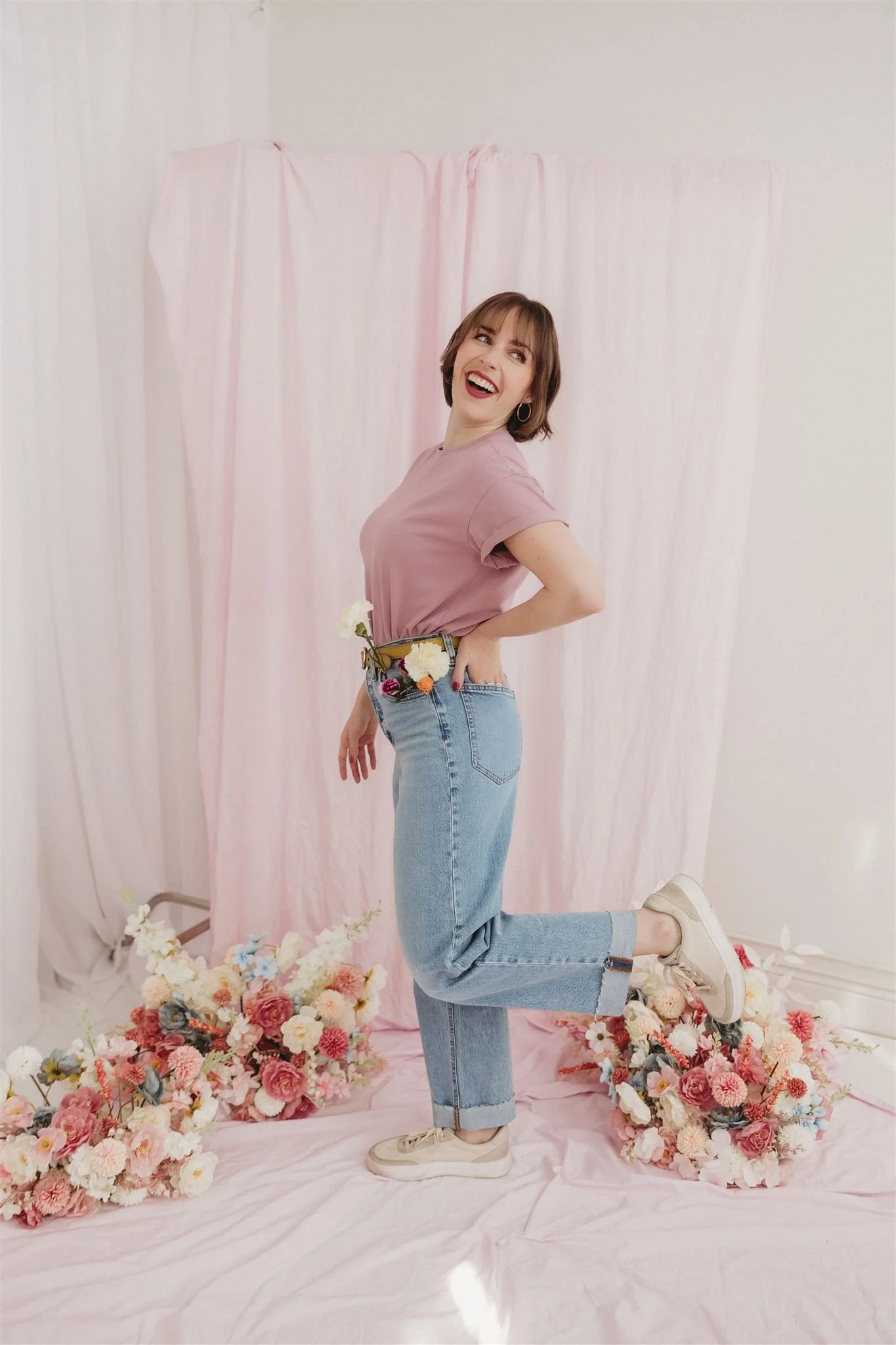 A woman with short brown hair, wearing a pink T-shirt, blue jeans, and white sneakers, standing among pink and white flowers, laughing and looking up, in front of a pink backdrop.