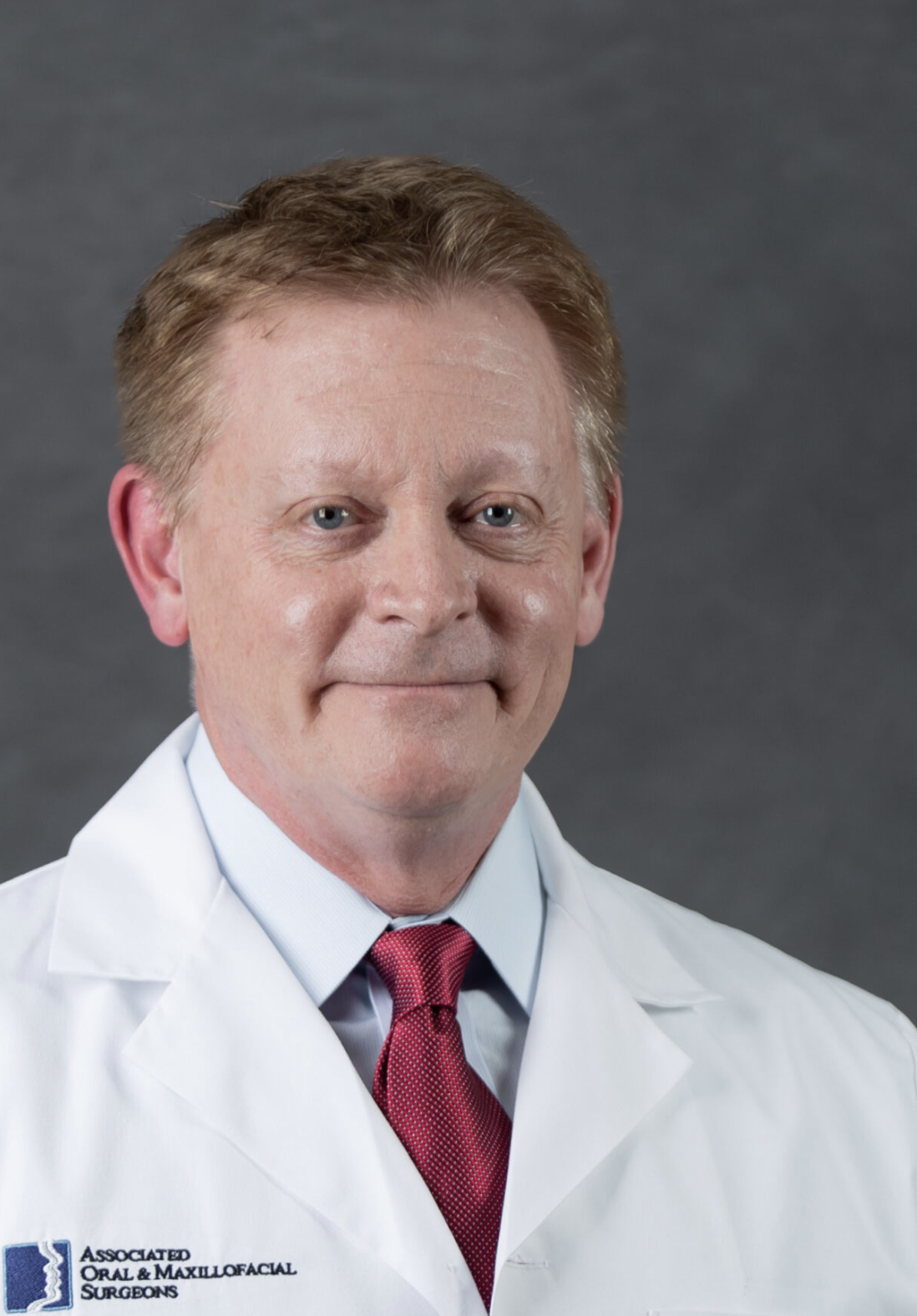 A male doctor wearing a white coat with a logo that says "Associated Oral & Maxillofacial Surgeons" and a red tie, standing against a gray background.
