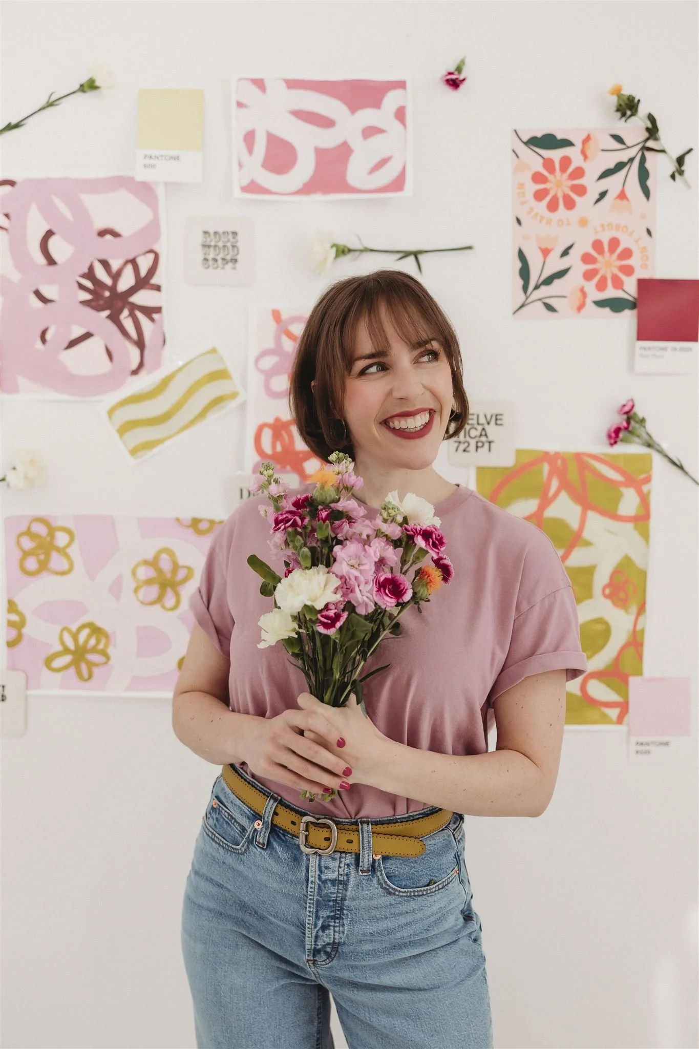 A woman holding a bouquet of pink and white flowers, smiling and looking to her left, in front of a wall decorated with various colorful abstract and floral patterned papers.