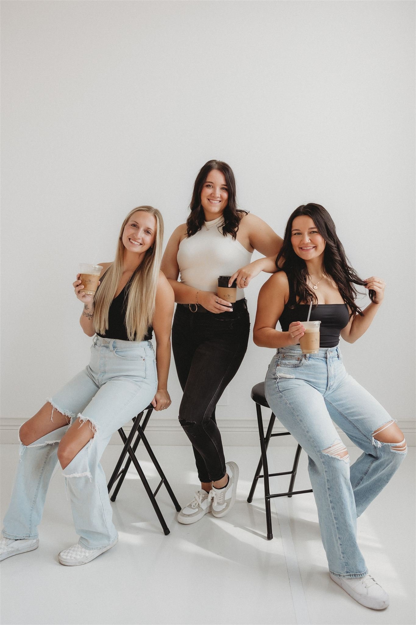 Three young women with long hair sit on black chairs and hold iced coffee drinks. They wear casual outfits with ripped jeans and black or white tops, smiling at the camera in a minimalistic room with white walls.