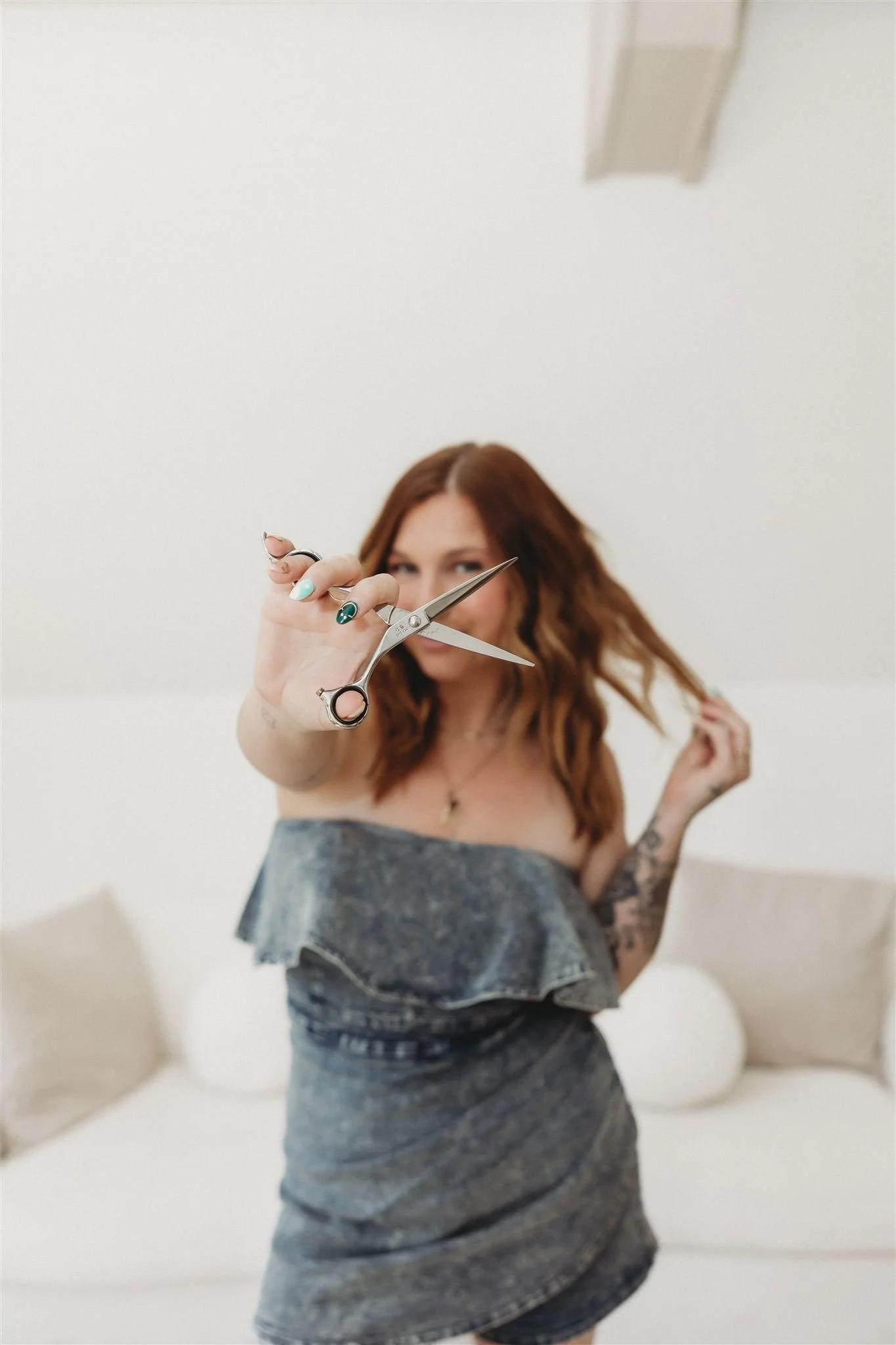A woman with red hair holding a pair of scissors towards the camera, standing in a brightly lit room with a white couch and pillows in the background.